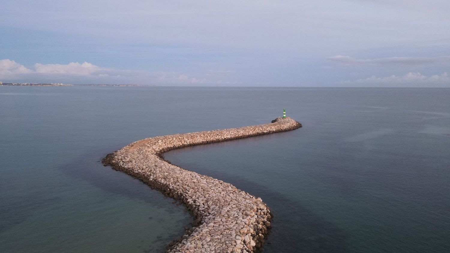 A curved stone breakwater jetty stretches into teal ocean waters under cloudy skies.