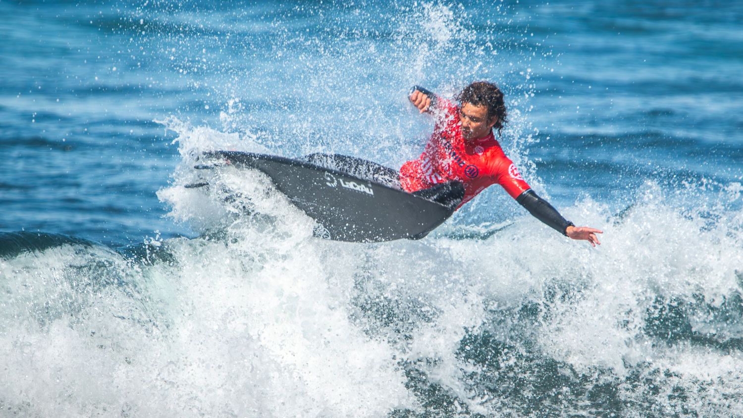 Surfer in red wetsuit performs dynamic aerial maneuver on breaking ocean wave with white spray splashing.