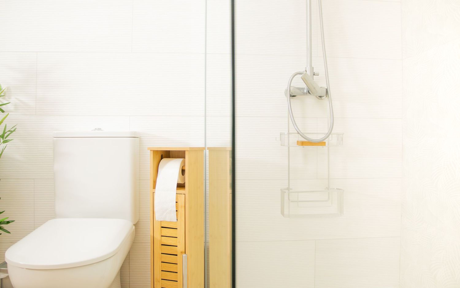 Modern minimalist bathroom interior featuring a white toilet, bamboo cabinet, and hung glass shower caddy.