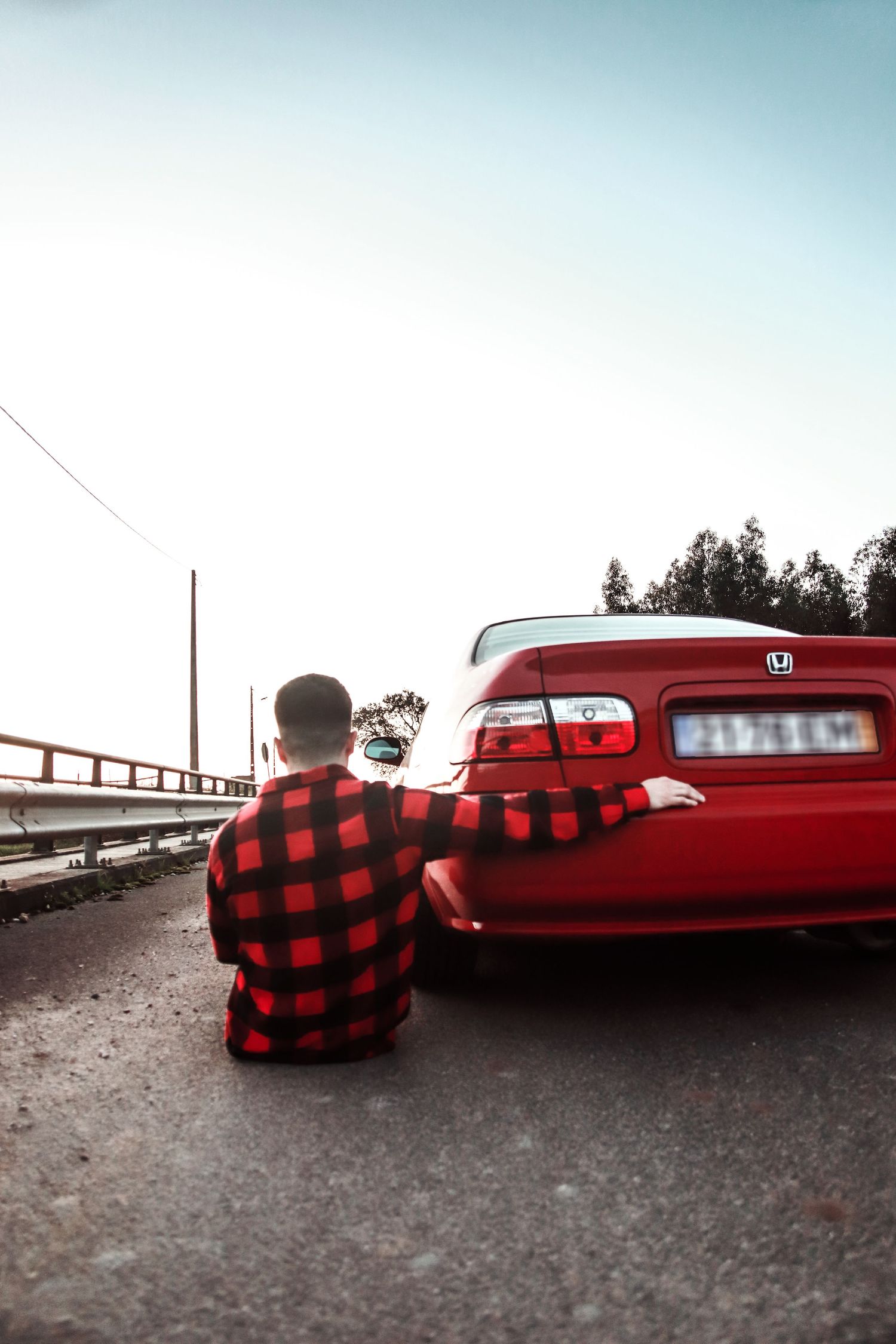Person in red plaid shirt sits on ground next to red Honda Civic at sunset.