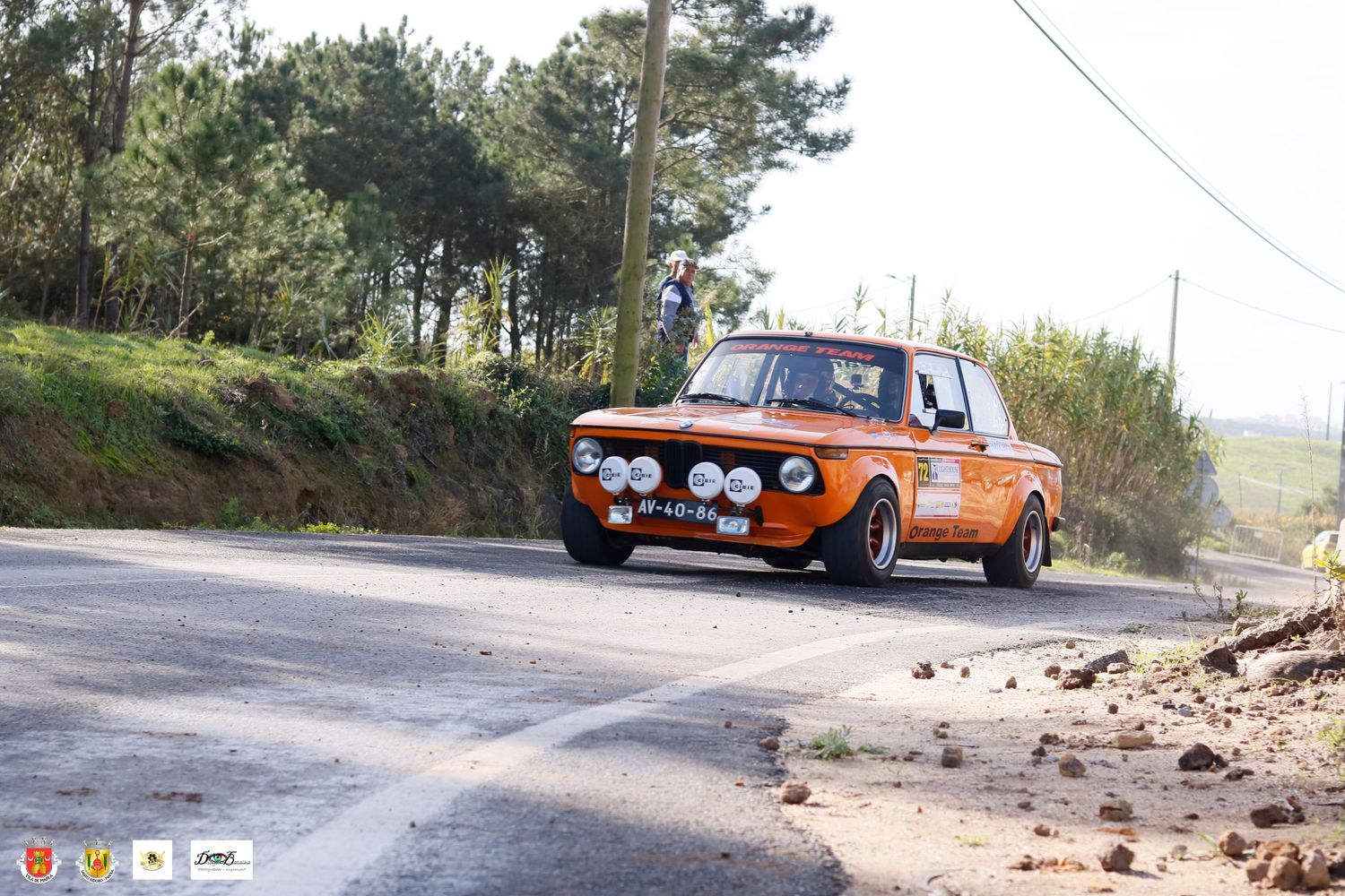 Vintage orange rally car jumping over crest on mountain road during racing competition.