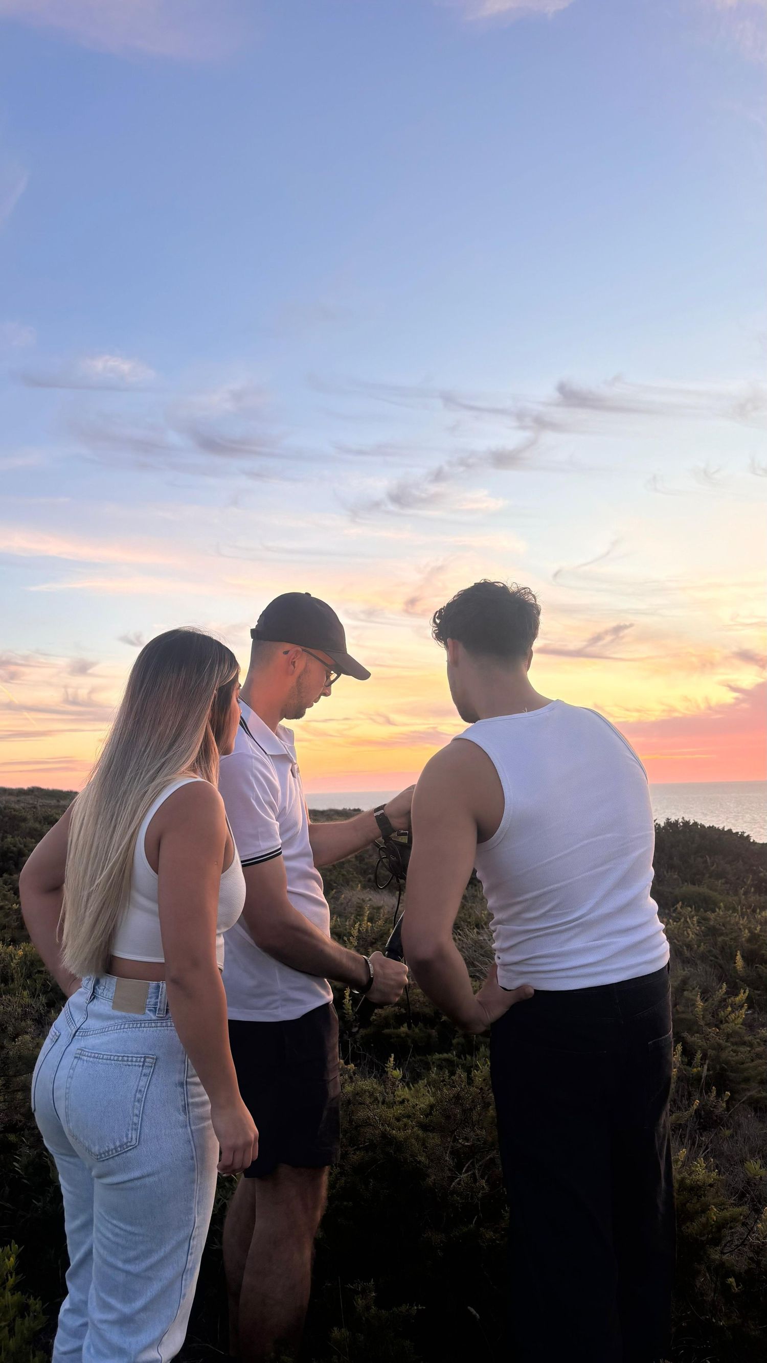 A group of friends stand together watching a colorful sunset over a scenic mountain landscape.
