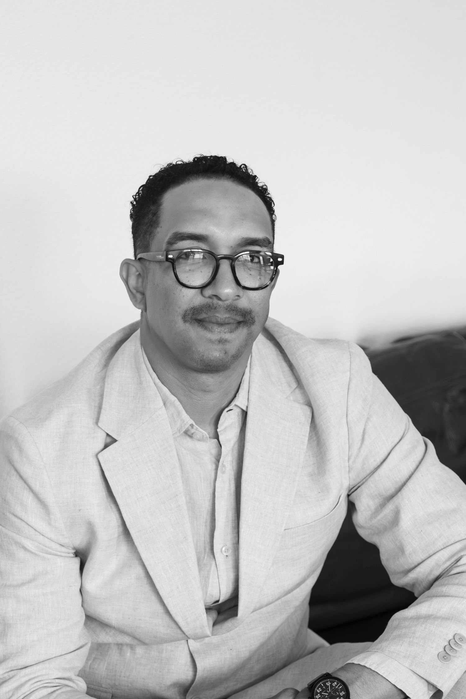 A man in a white suit jacket and glasses smiles at the camera while reclining in a casual pose.