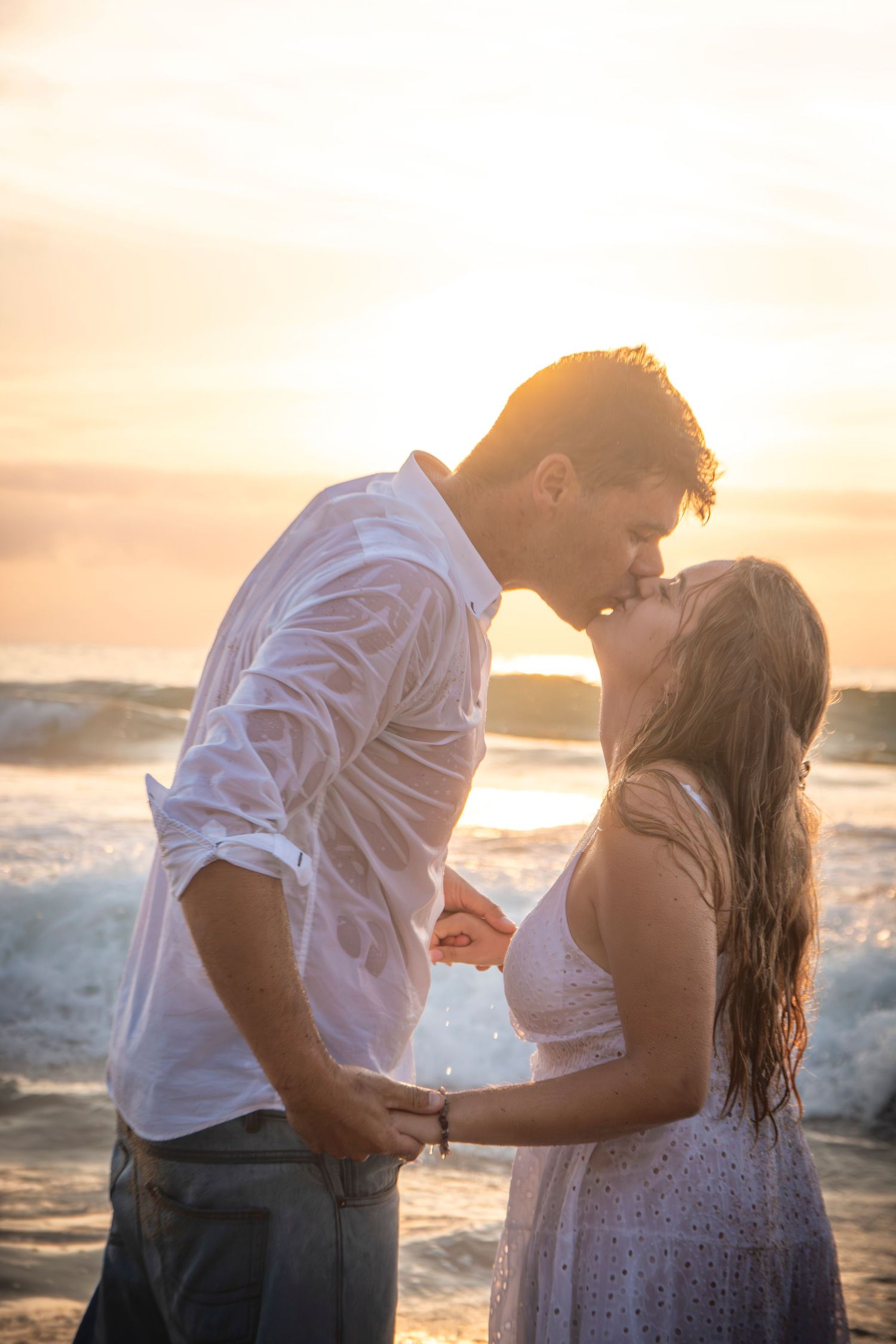 A romantic couple embraces at sunset by the ocean beach while golden light illuminates their silhouettes.
