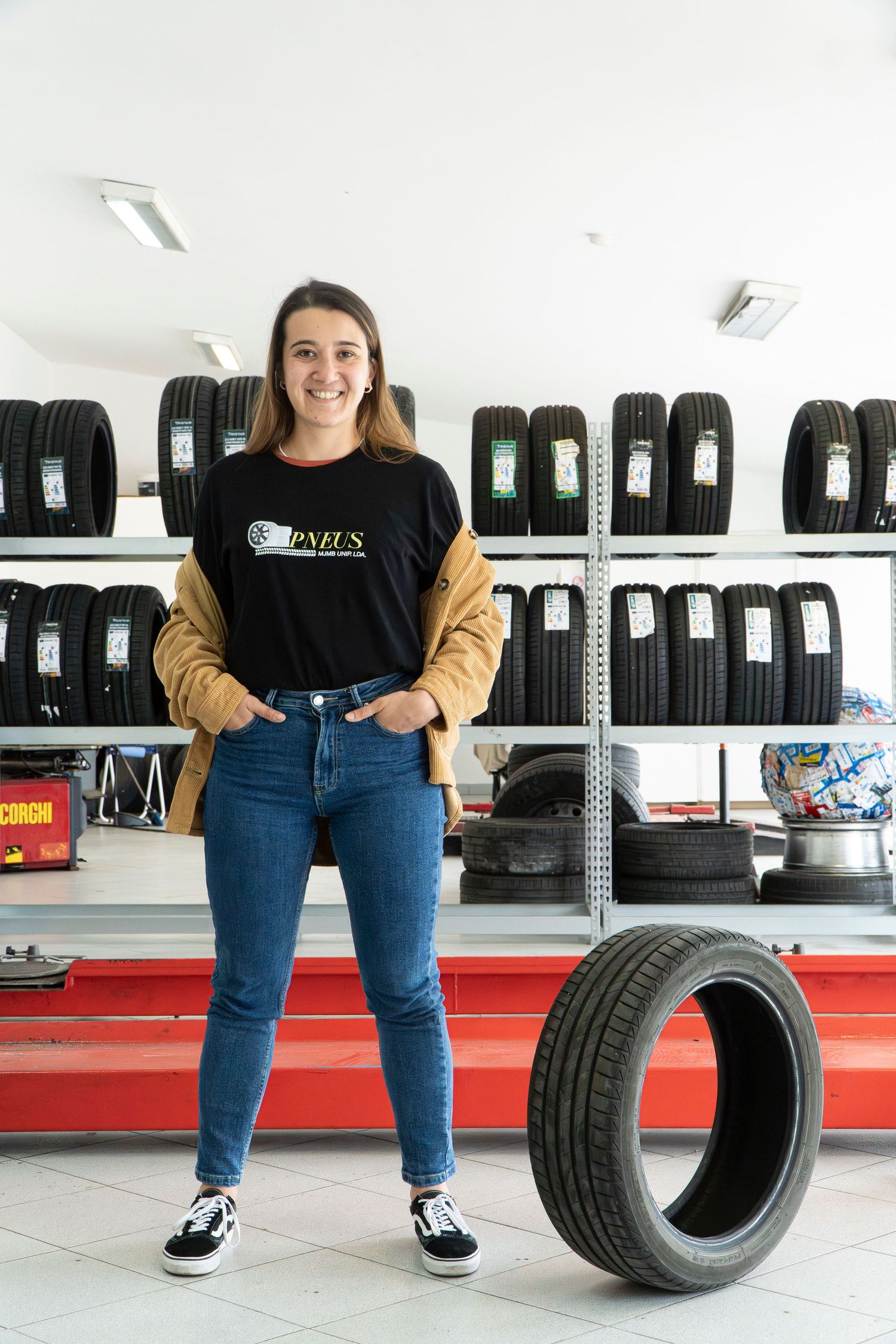 Young woman in a Pneus t-shirt smiling in a tire shop, standing beside a car tire with shelves of tires behind her.