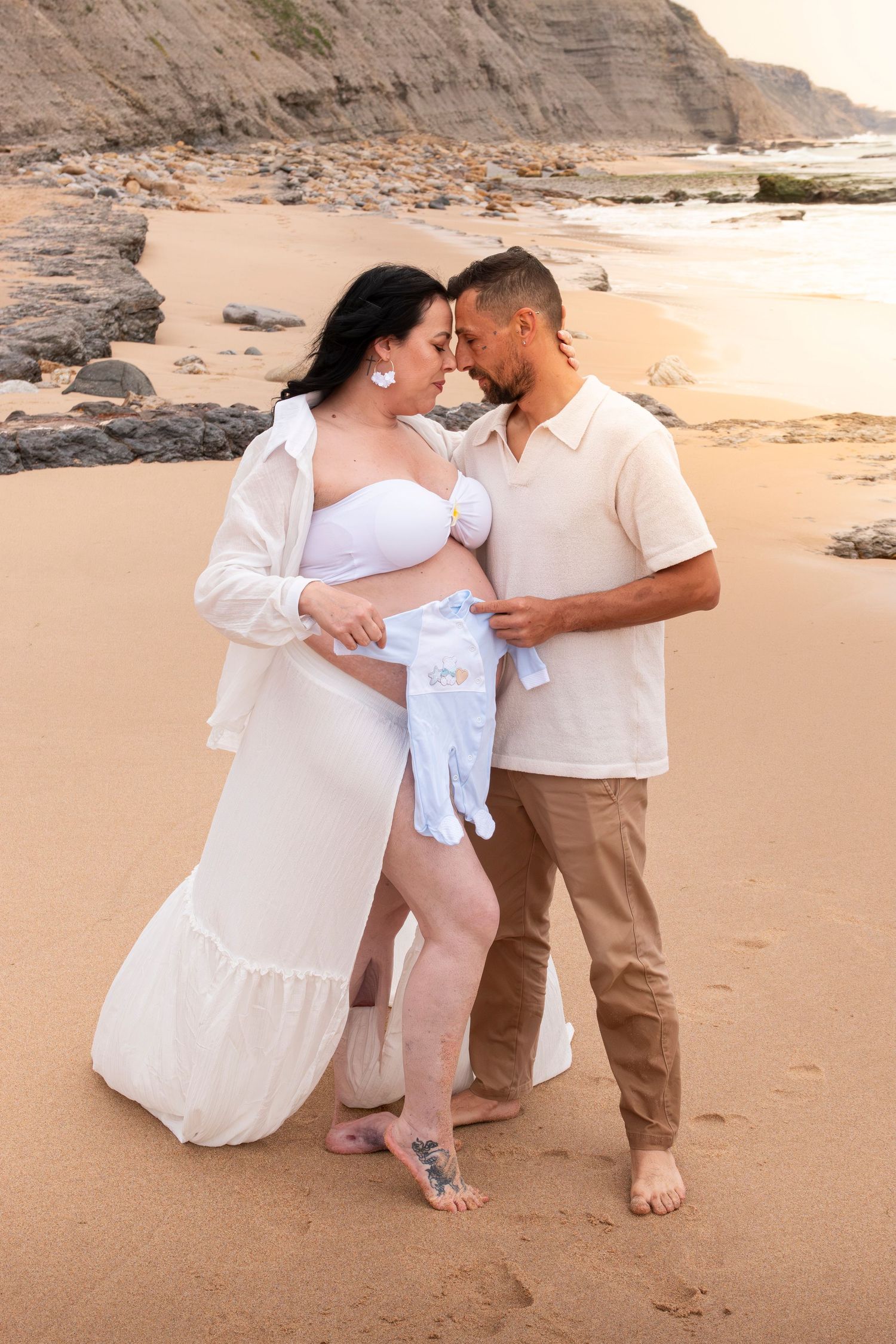 Pregnant couple sharing a tender moment on a sandy beach at sunset, holding baby clothes near rocky cliffs.