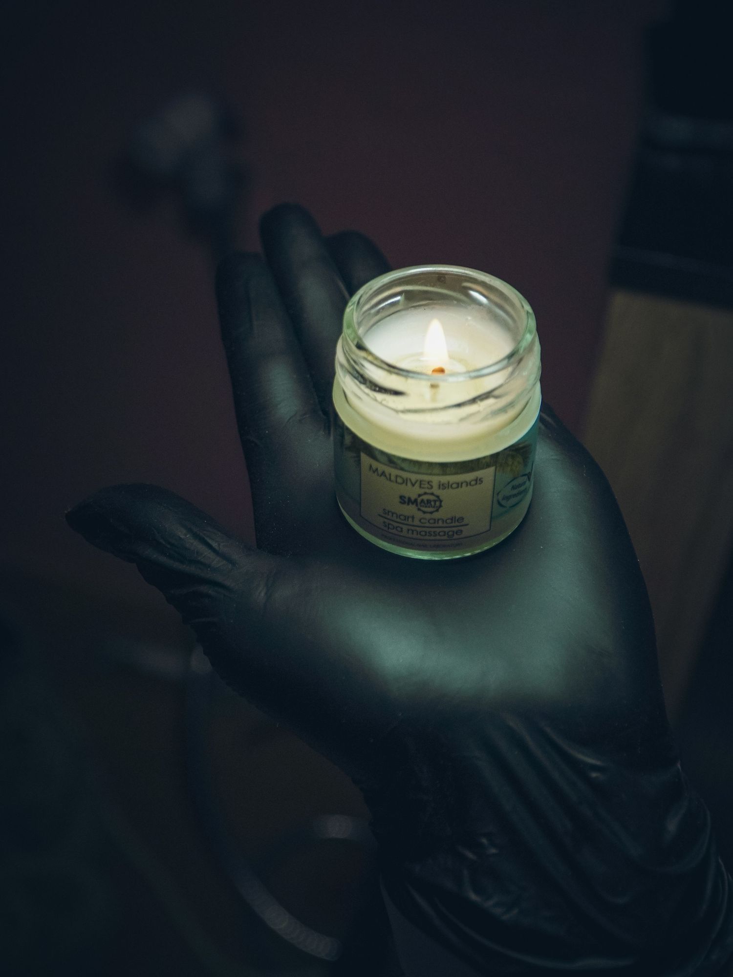 A lit candle in a small jar held by a hand wearing black latex gloves against a dark background.