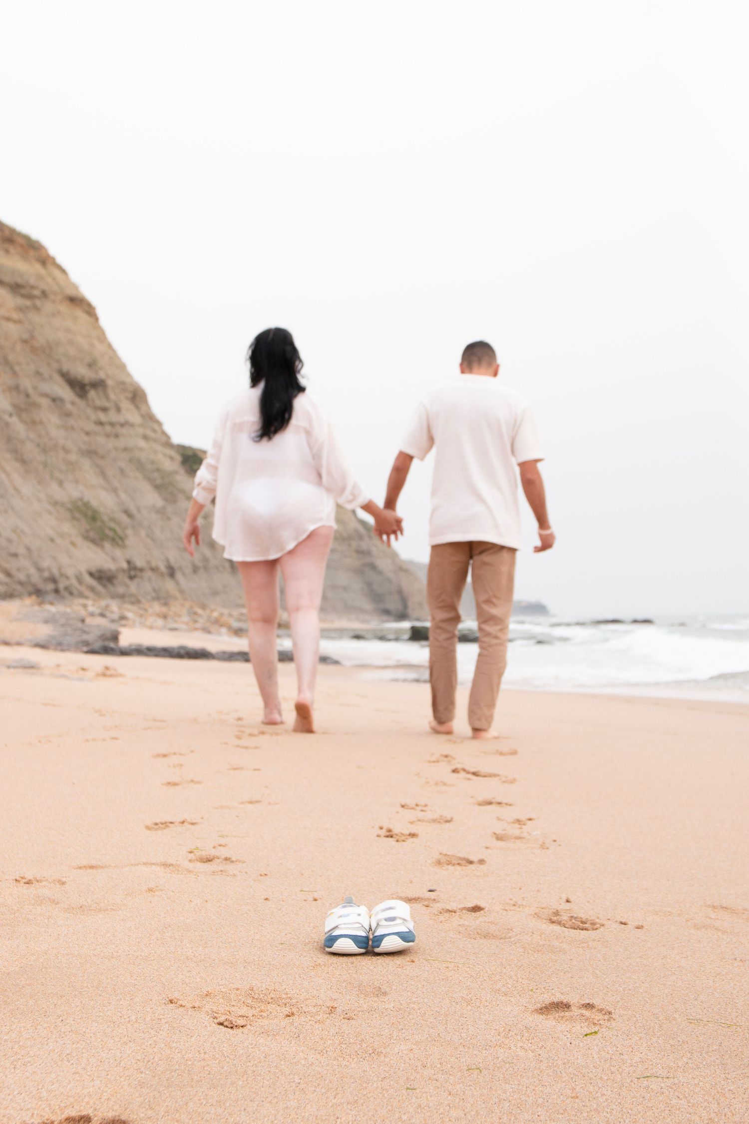 Pregnant couple walking hand in hand on sandy beach, baby shoes in foreground, ocean and cliffs in background.