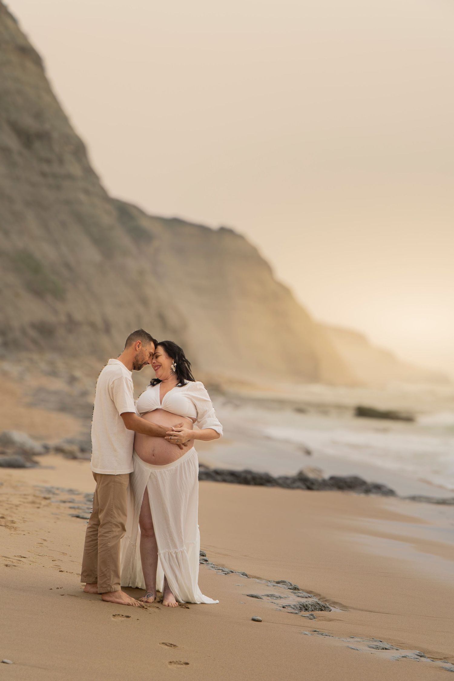 Couple sharing tender moment on sandy beach at sunset, pregnant woman in white dress cradled by partner near coastal cliffs.
