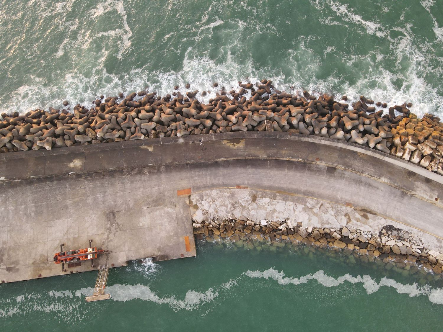 Aerial view of sea lions lounging on a curved concrete breakwater with crashing waves in turquoise ocean waters.