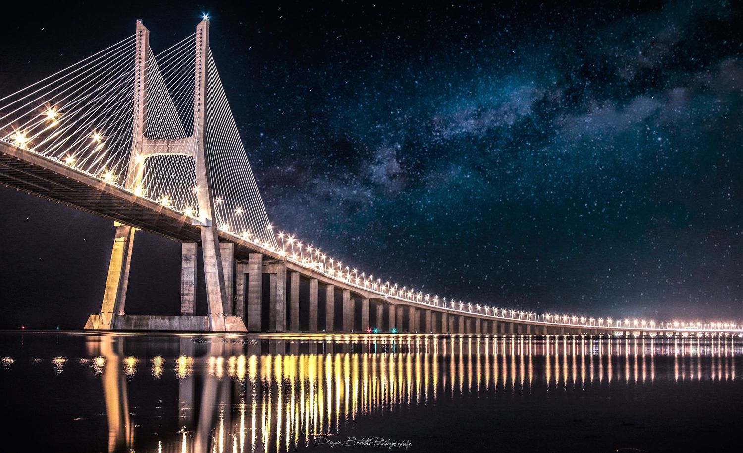 Long illuminated bridge stretches across dark water beneath a starry night sky, reflecting golden lights on calm surface.