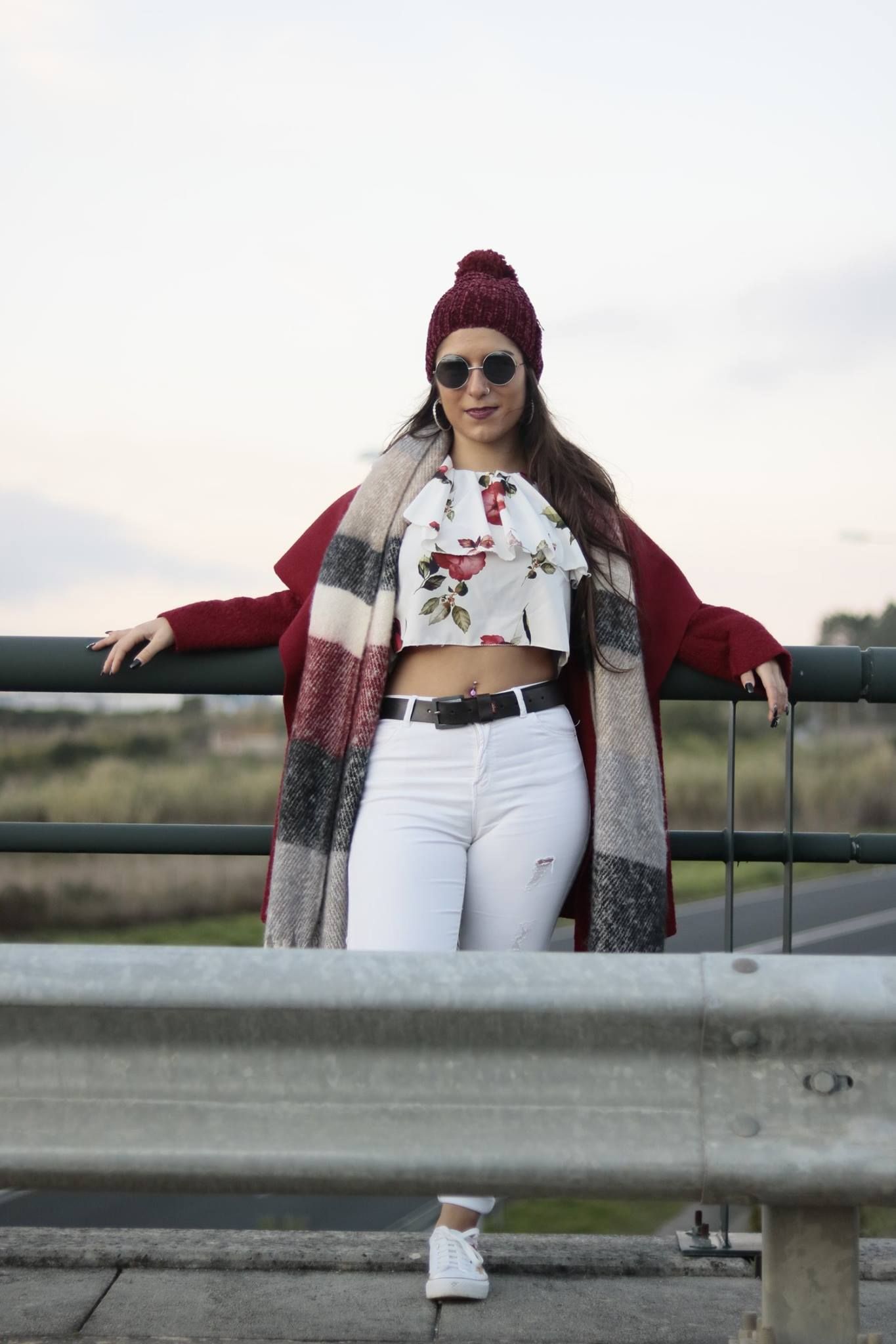 Fashionable winter outfit with red beanie, floral crop top and striped cardigan against rural backdrop.