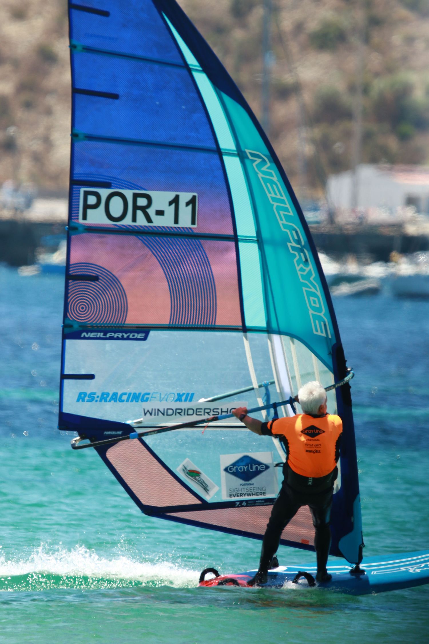 Windsurfer with blue and teal POR-11 sail maneuvering in coastal waters.