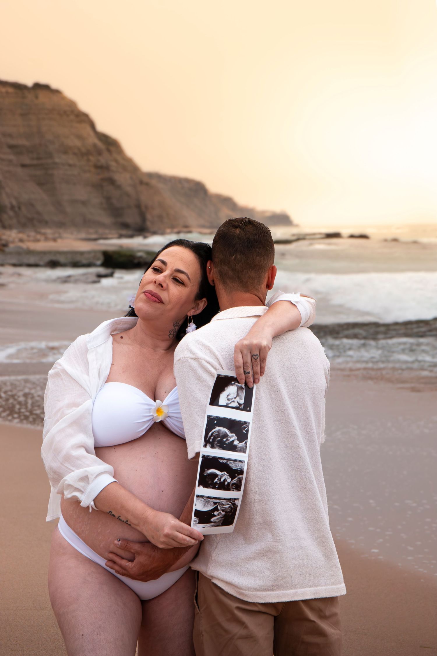 Pregnant woman in white bikini holds ultrasound photos while embracing partner on beach at sunset.