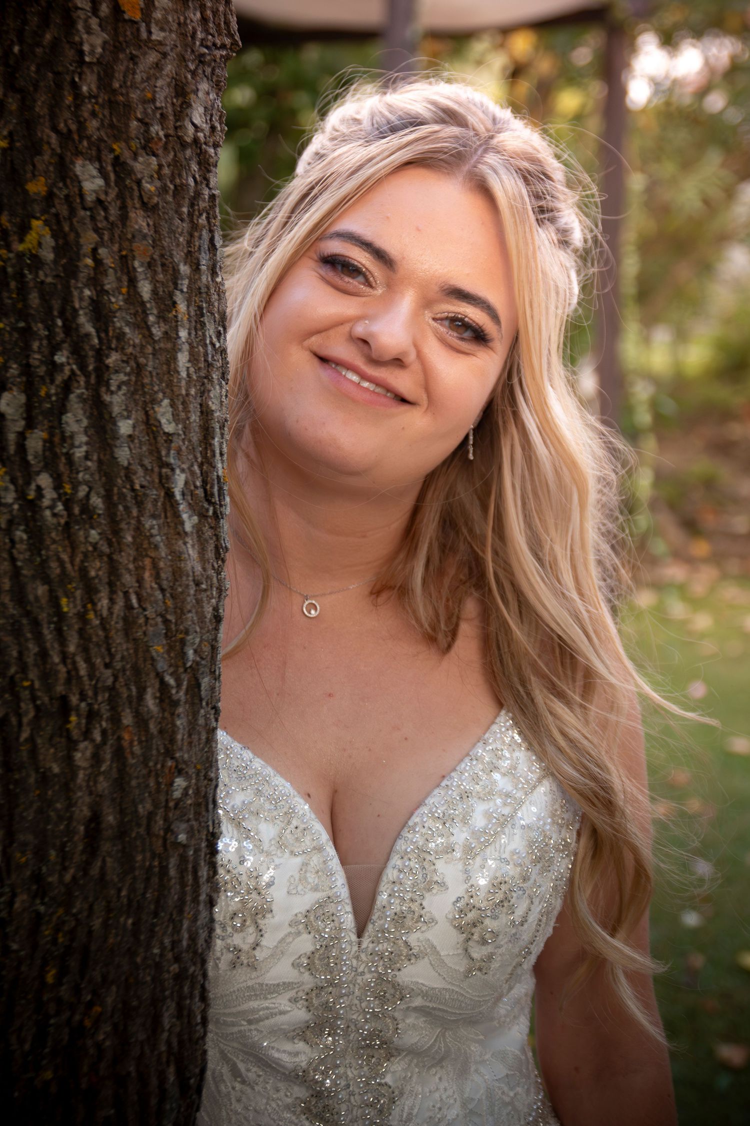 Smiling blonde bride in a beaded white wedding gown leaning against a tree in an outdoor garden setting.