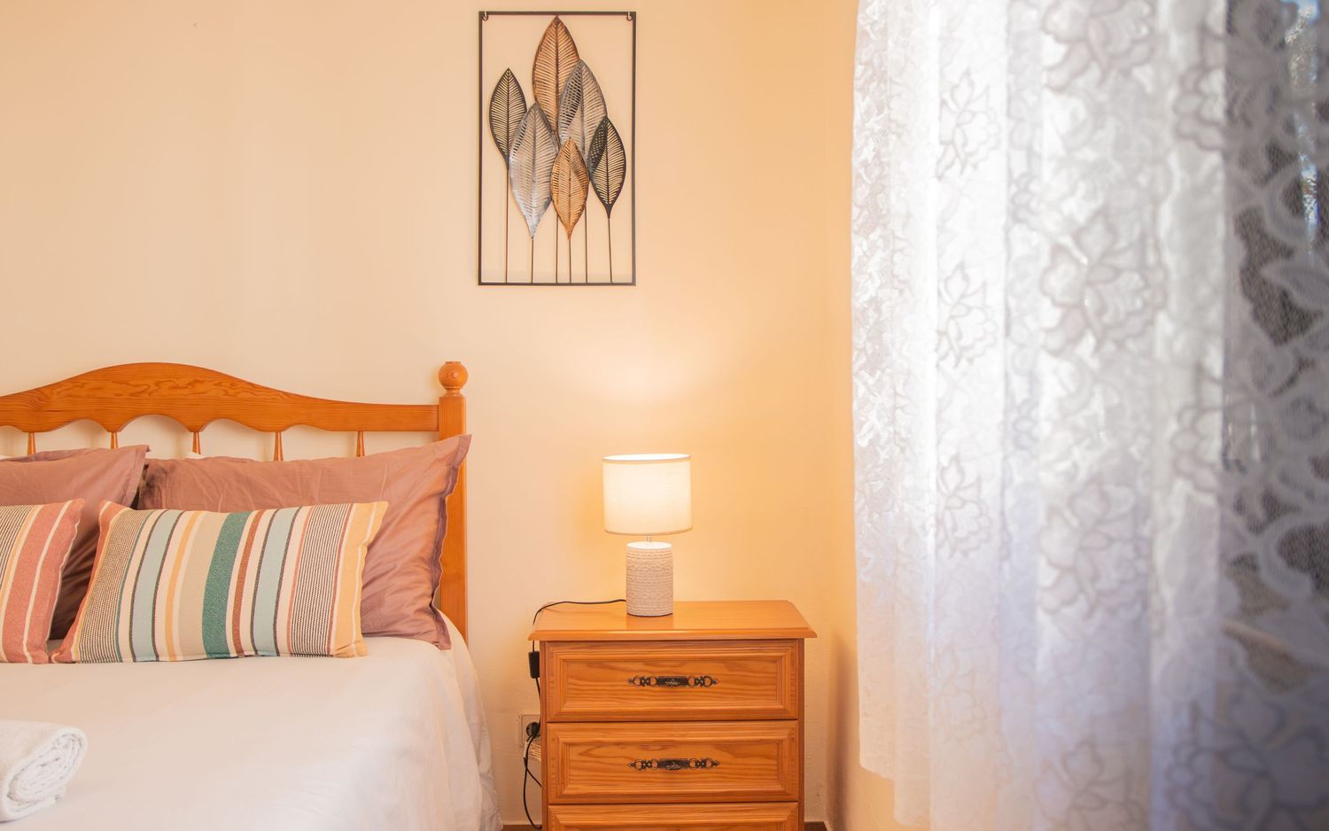 Cozy bedroom interior with wooden headboard, striped pillows, nightstand, table lamp and floral wall art.
