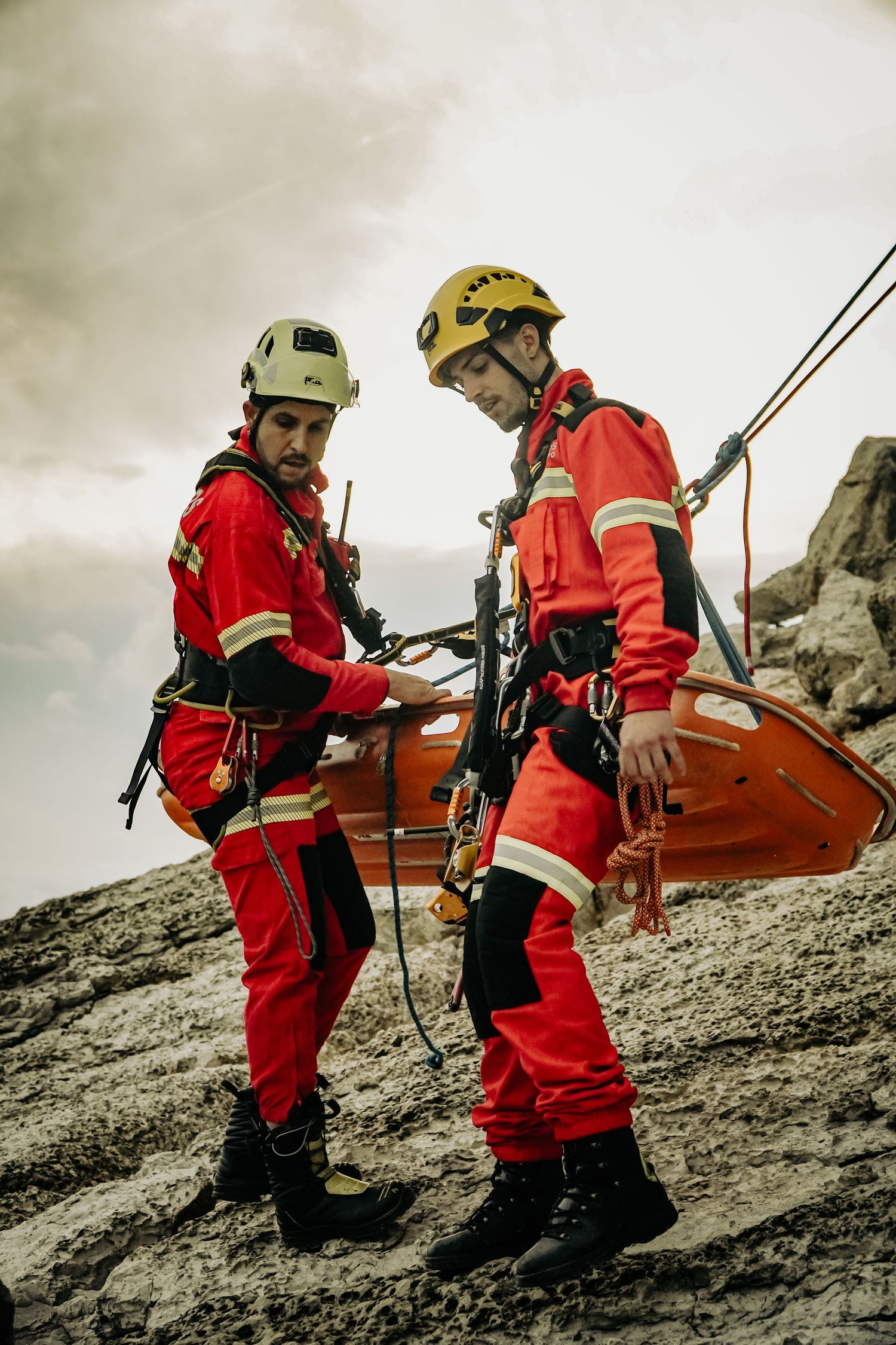 Two rescue workers in red safety gear with climbing equipment prepare for mountain operations on rocky terrain.