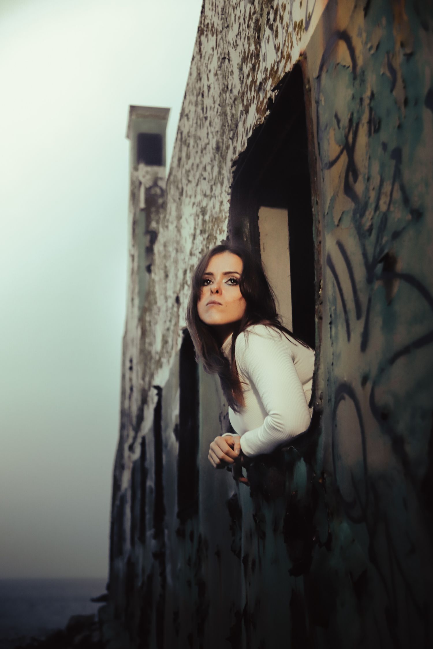 A person in a white sweater leans out from a dark opening in an old concrete structure with graffiti.