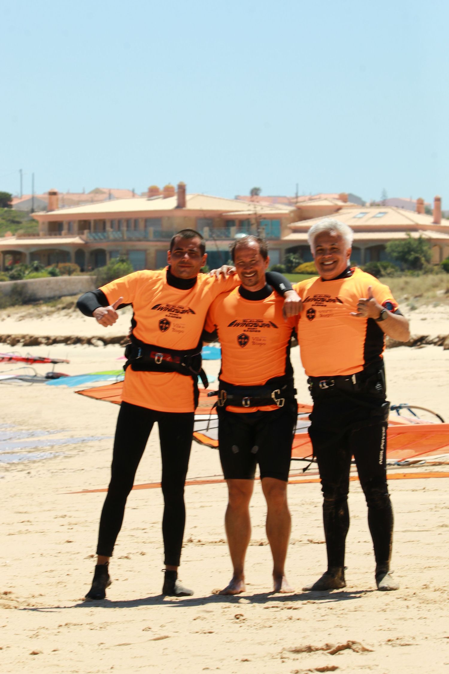 Three people in matching orange shirts and wetsuits pose on a beach with windsurfing equipment.