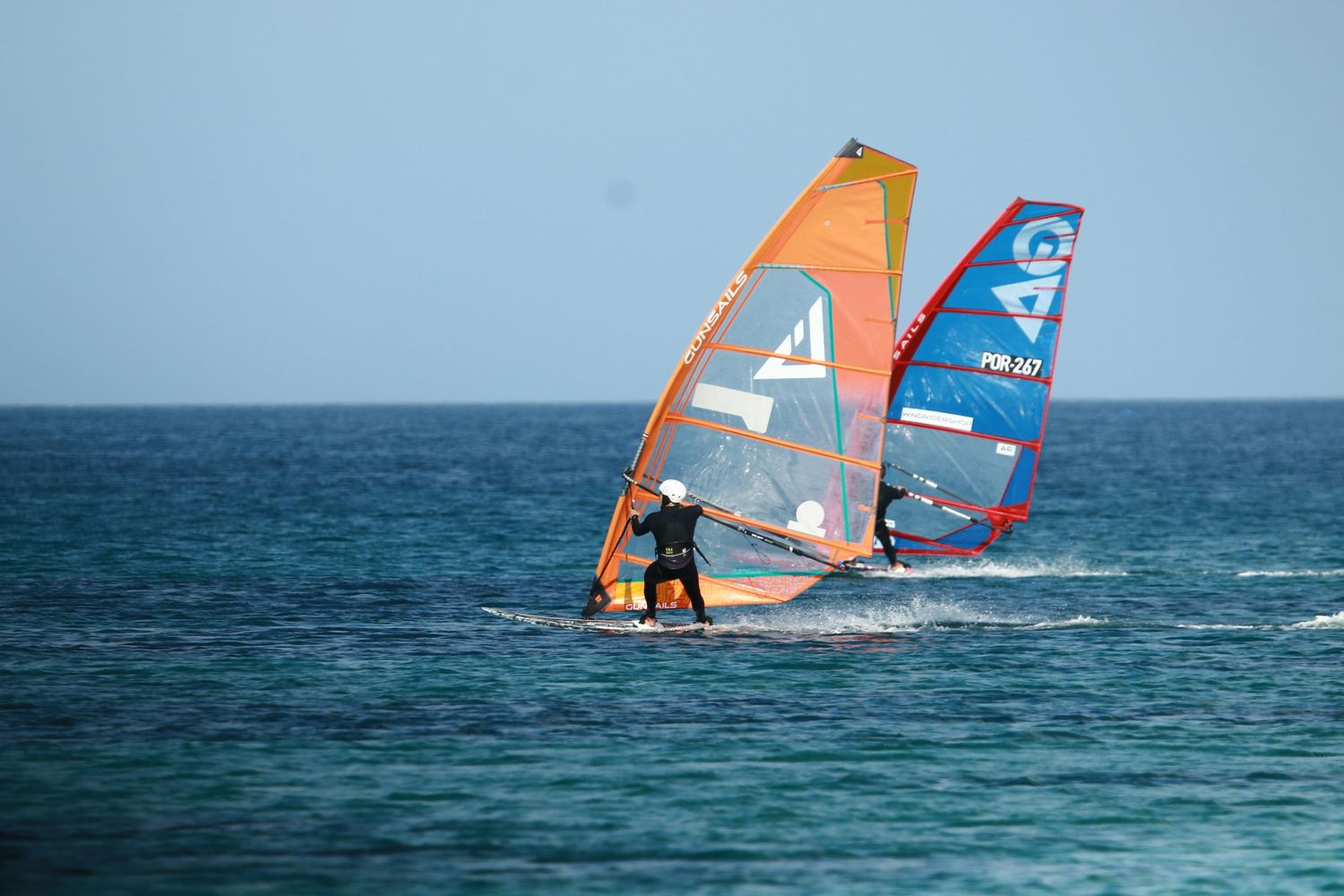 Two windsurfers with colorful sails practicing on calm ocean waters.