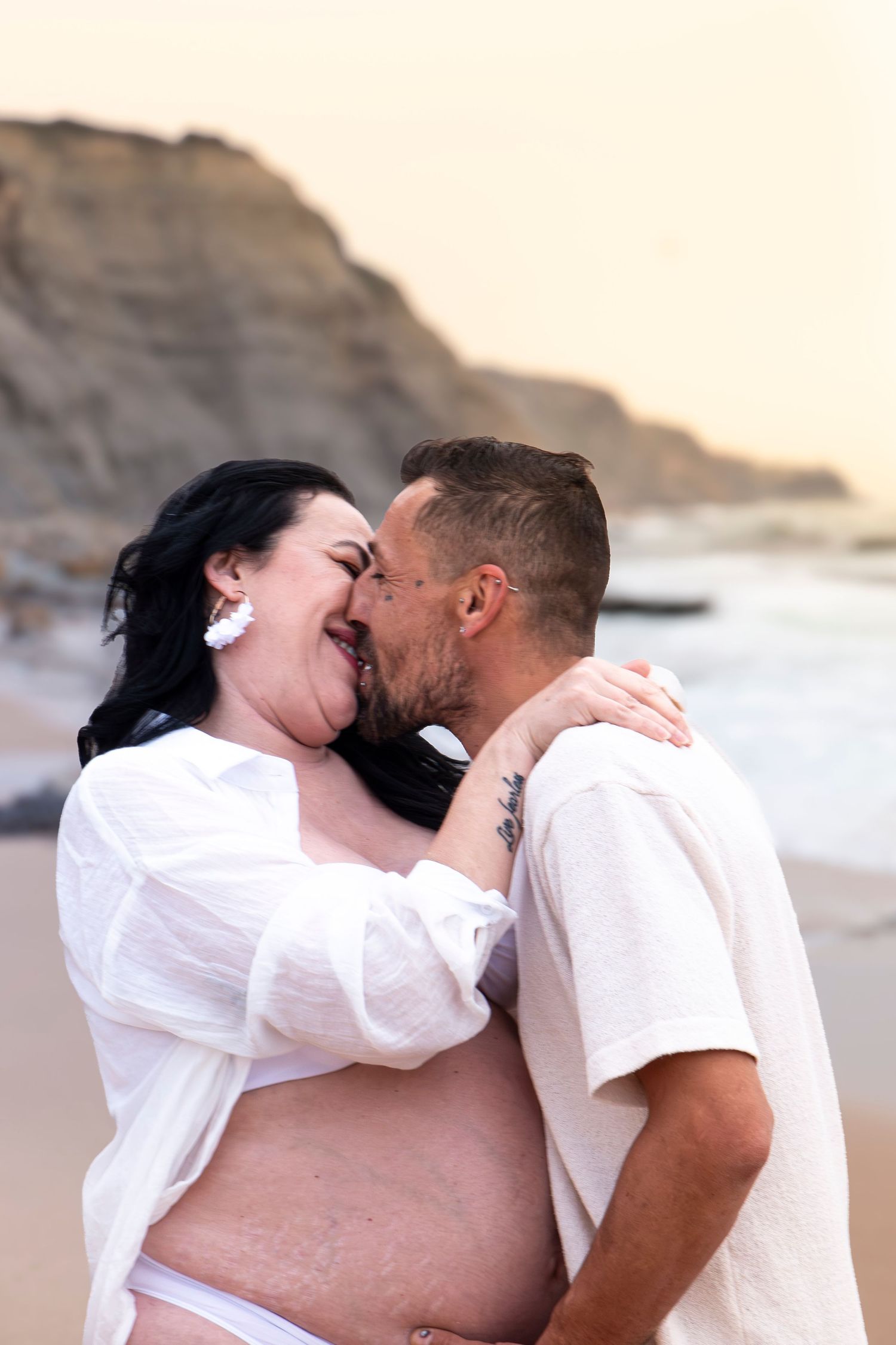 Pregnant couple sharing a tender kiss on a beach, with rocky cliffs and ocean waves in the background.