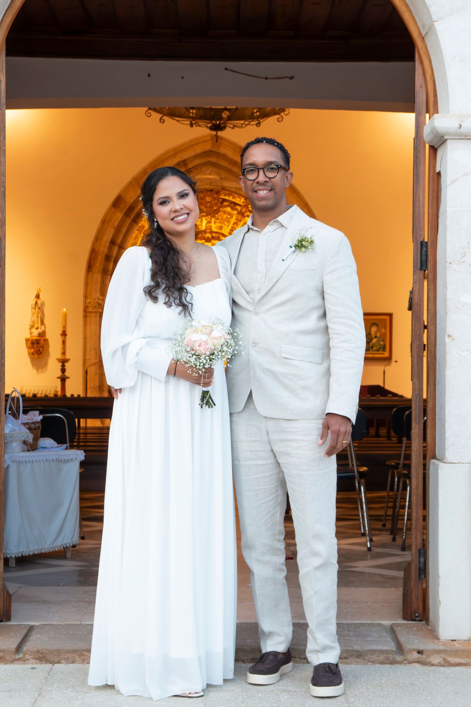A couple poses together in white formal attire against arched doorways at a wedding celebration.