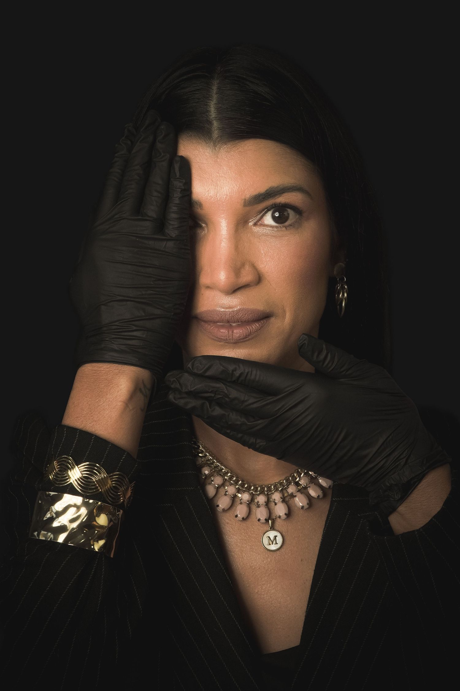 Dramatic portrait with black gloves, gold bangle and ornate necklace against dark background.