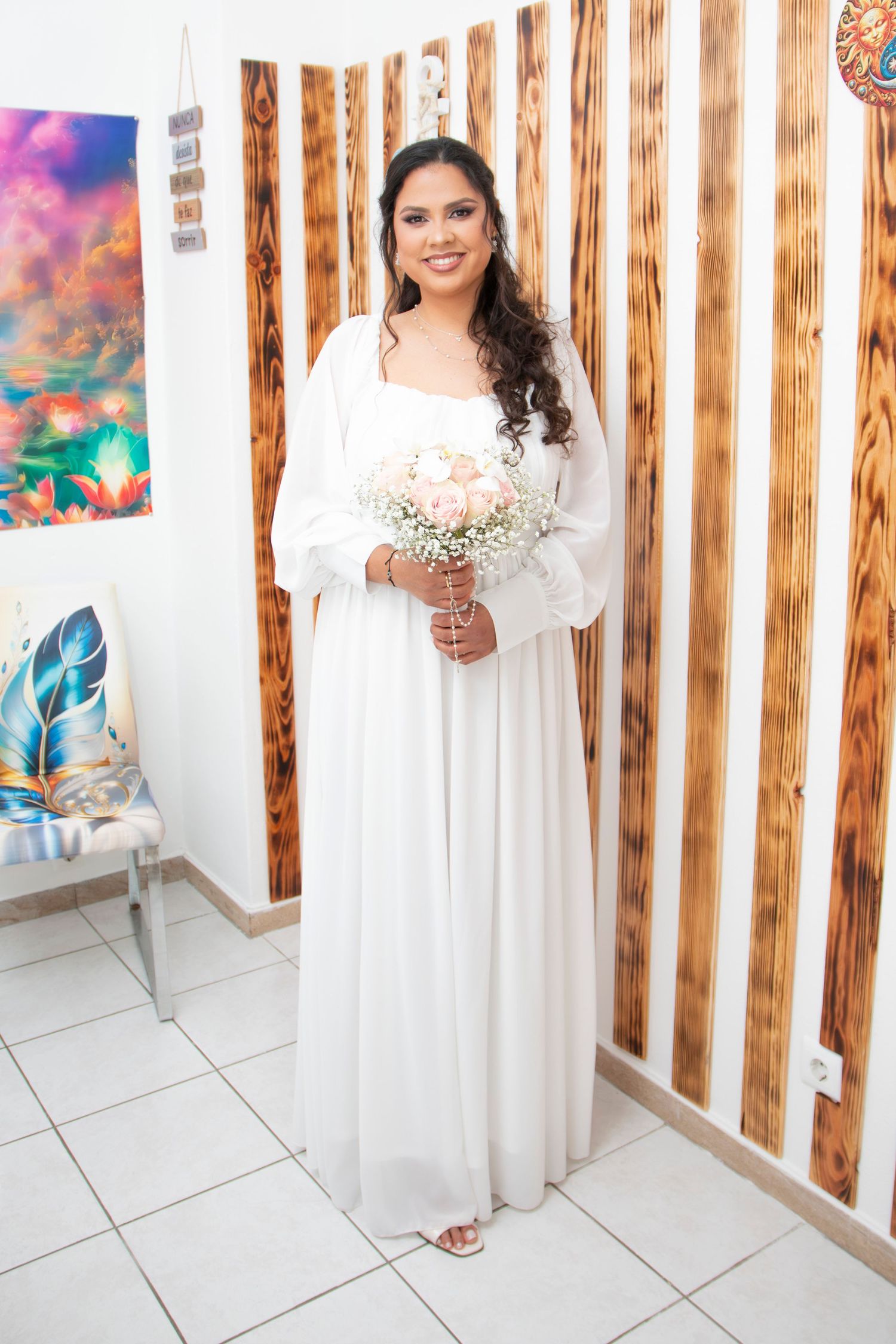 A person in a flowing white gown poses elegantly in a rustic wooden-walled room decorated with colorful artwork.