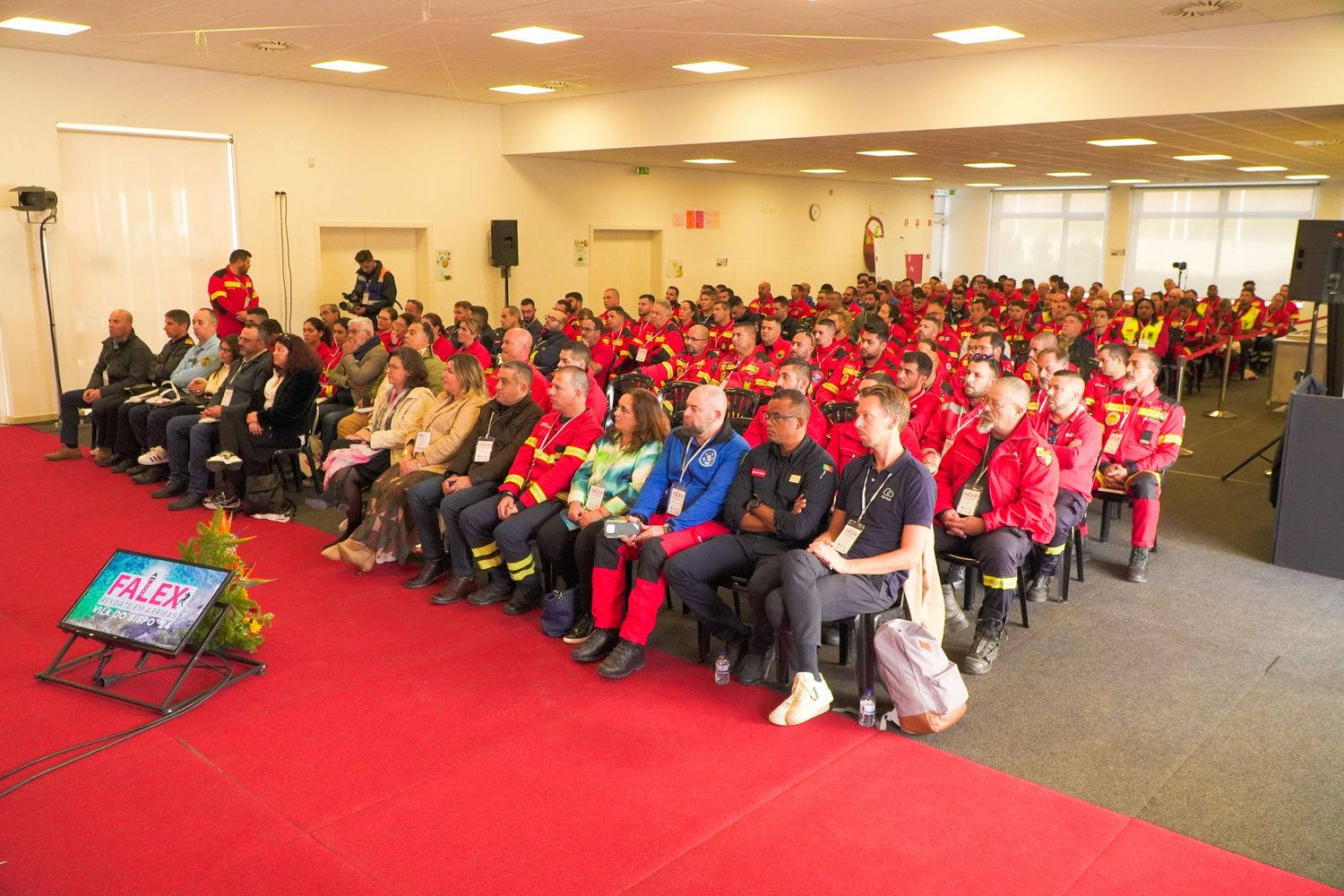 Large audience wearing red shirts seated in a modern conference room.