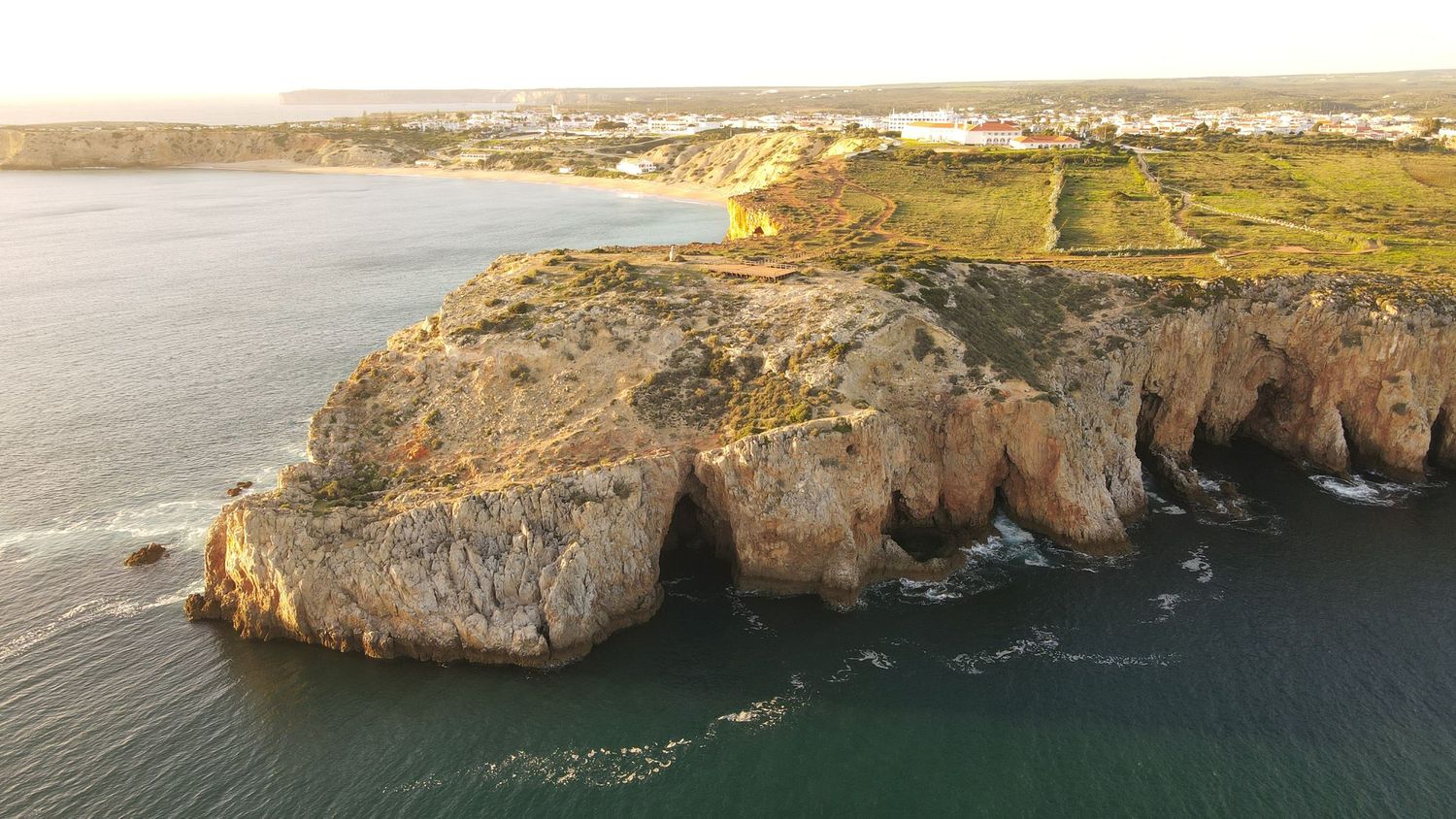 Dramatic aerial view of rugged coastal cliffs along the Portuguese coastline with farmland visible in the distance.
