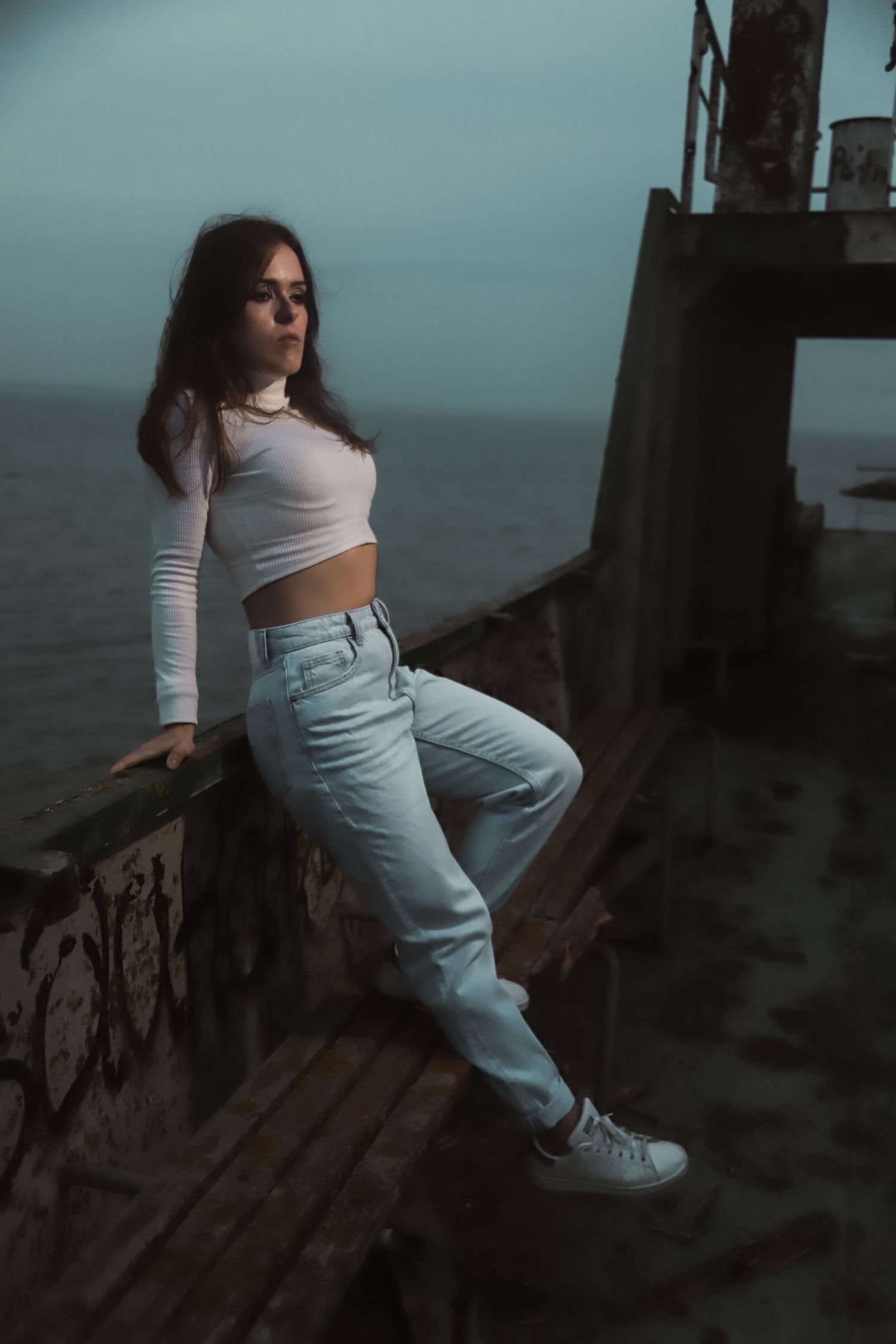 A moody portrait of someone in a white crop top and light jeans leaning against a railing by the ocean at dusk.