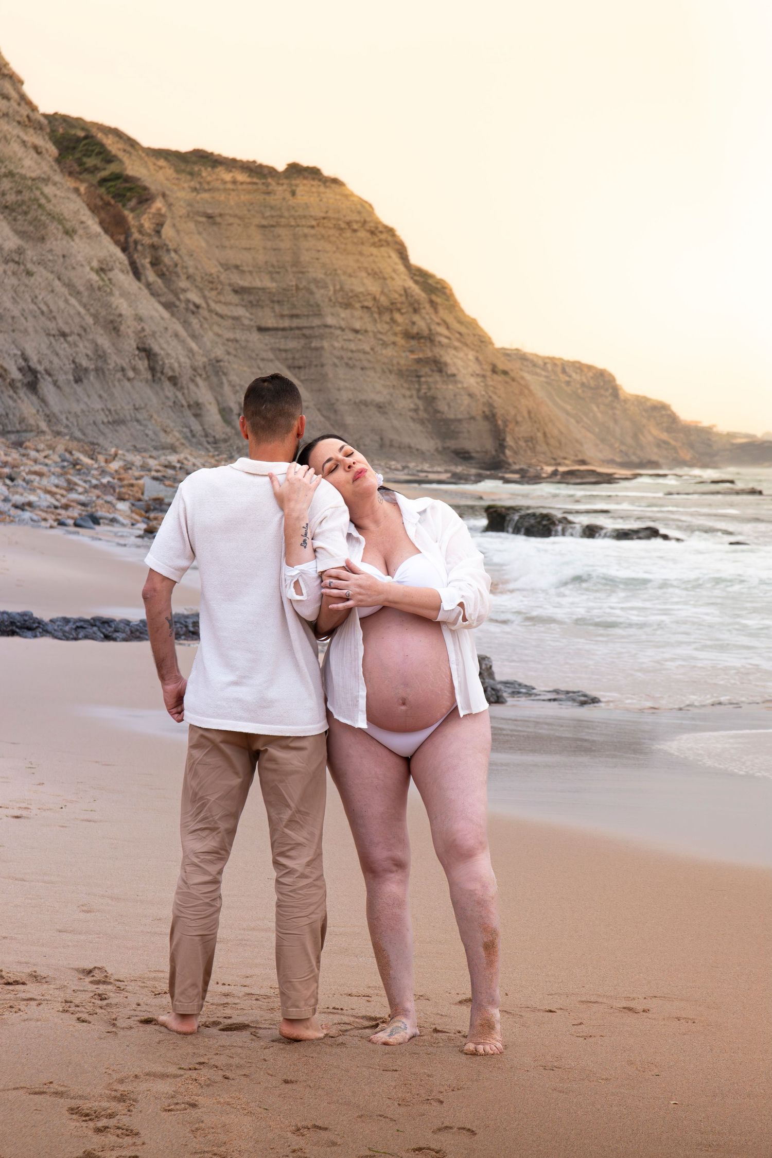 Pregnant couple embracing on sandy beach at sunset with rocky cliffs and ocean waves in background.