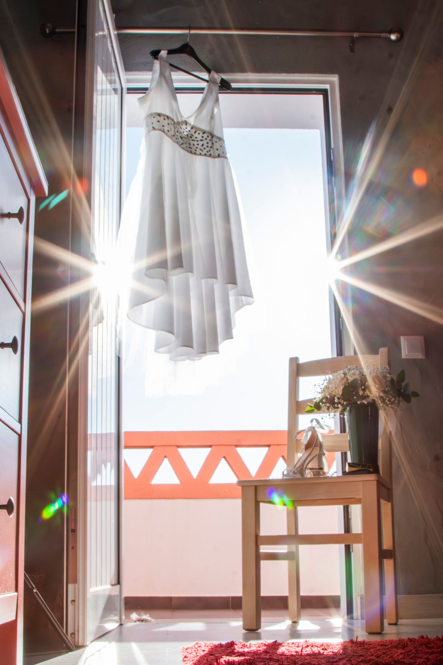 White wedding gown with beaded details hanging in doorway with sunlight creating lens flares.