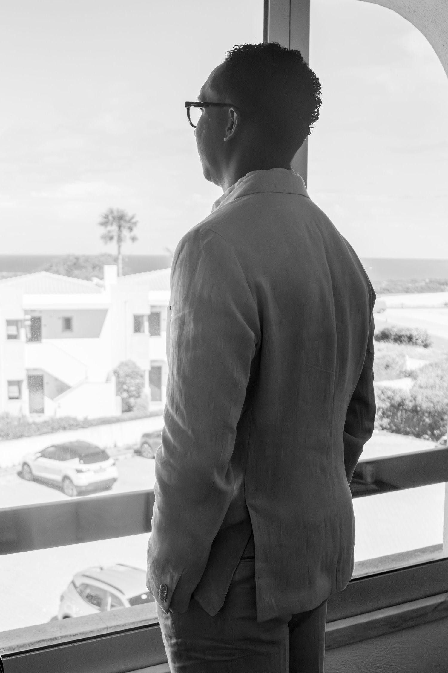 Man in dark clothing stands at window looking out across an urban landscape in black and white photography.