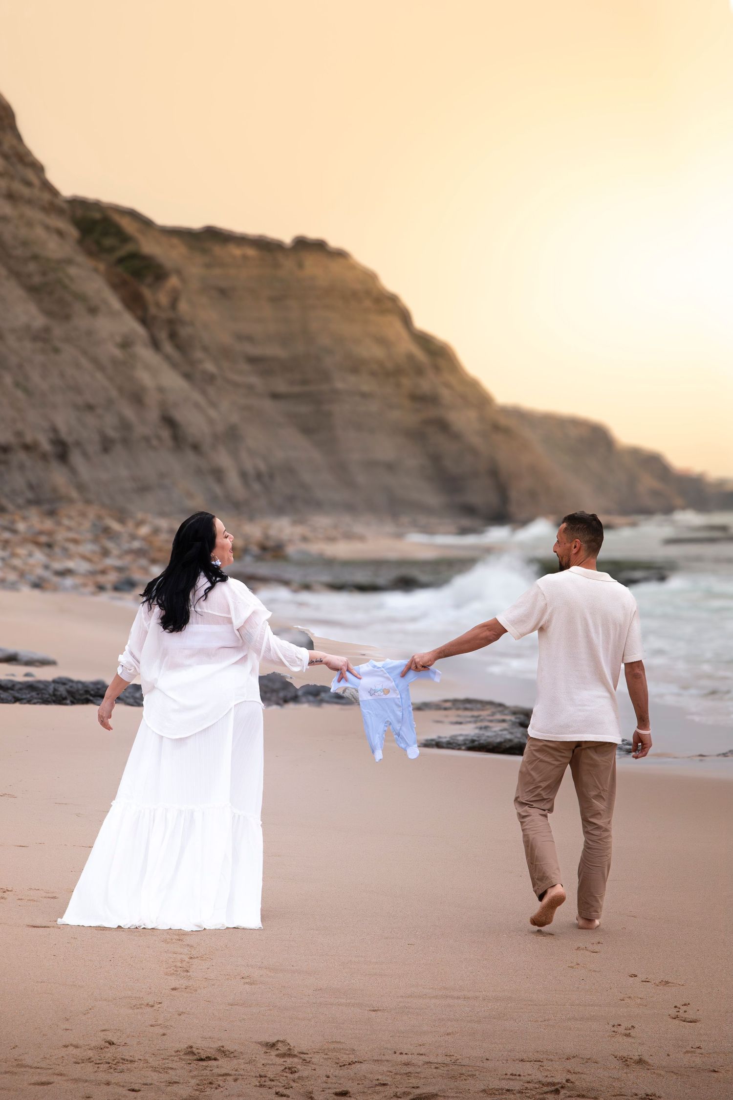 Couple walking on beach at sunset holding baby clothes, with cliffs and ocean waves in the background.