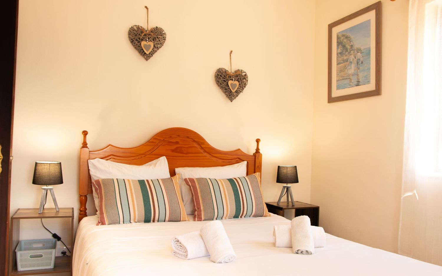Cozy bedroom with wooden headboard bed, striped pillows, bedside lamps and decorative heart ornaments on cream walls.