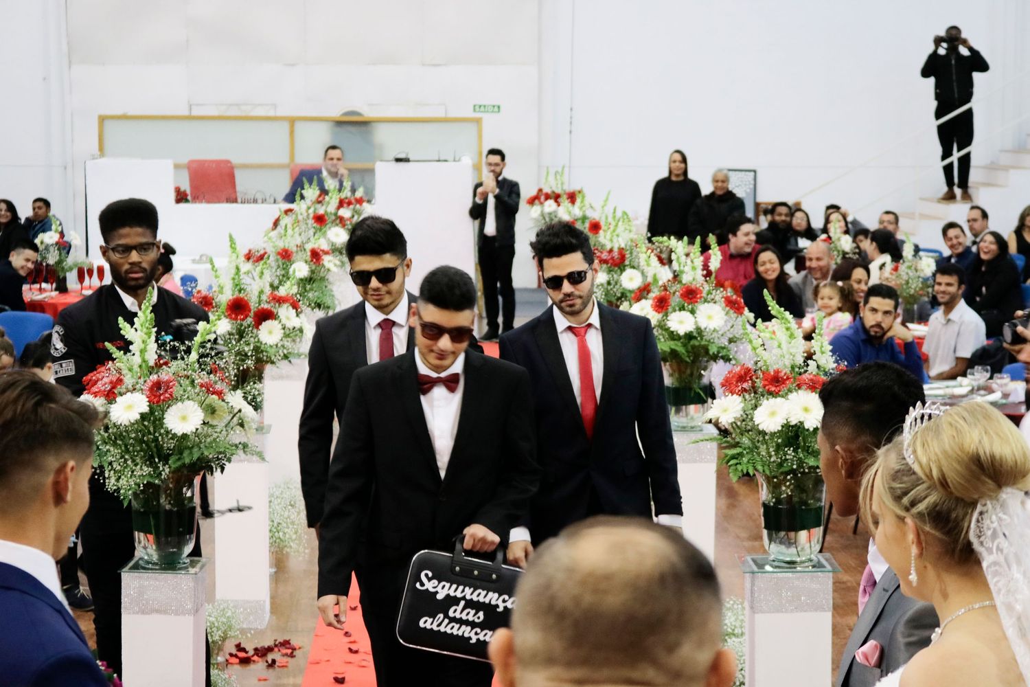 Groomsmen in dark suits walk down aisle decorated with red and white floral arrangements.