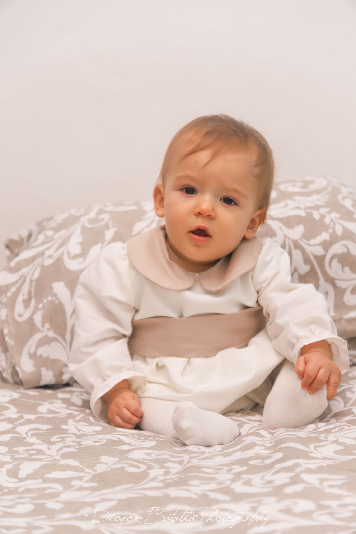 Baby in white christening gown lying on decorative white bedding.