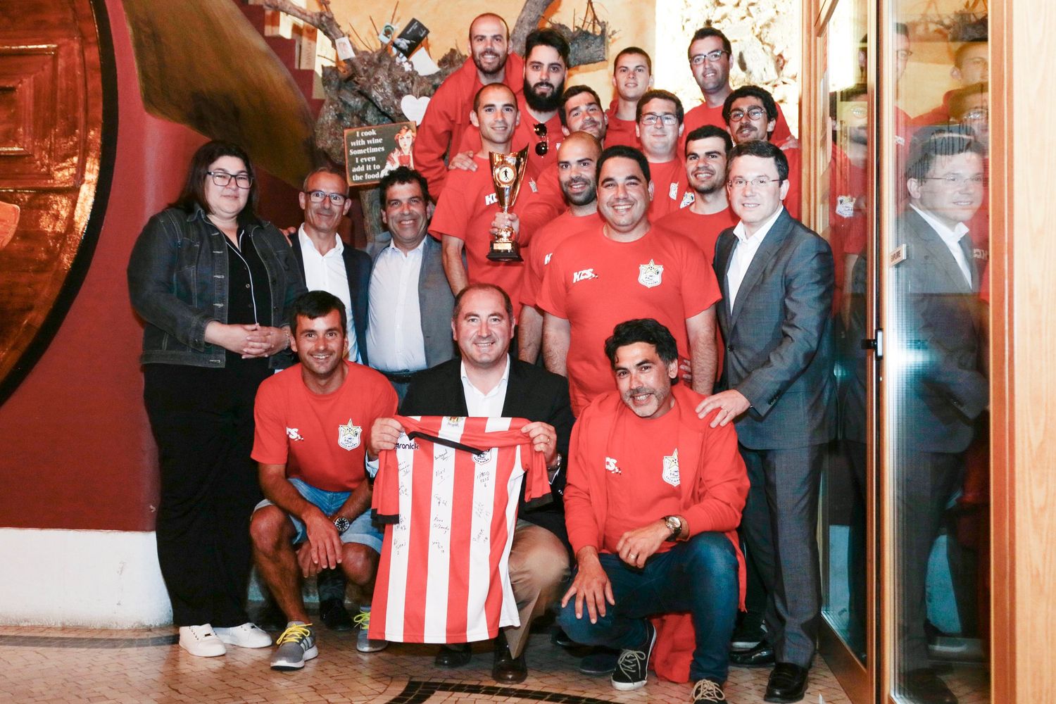A sports team in red jerseys poses for a group photo with a trophy and a striped jersey displayed.