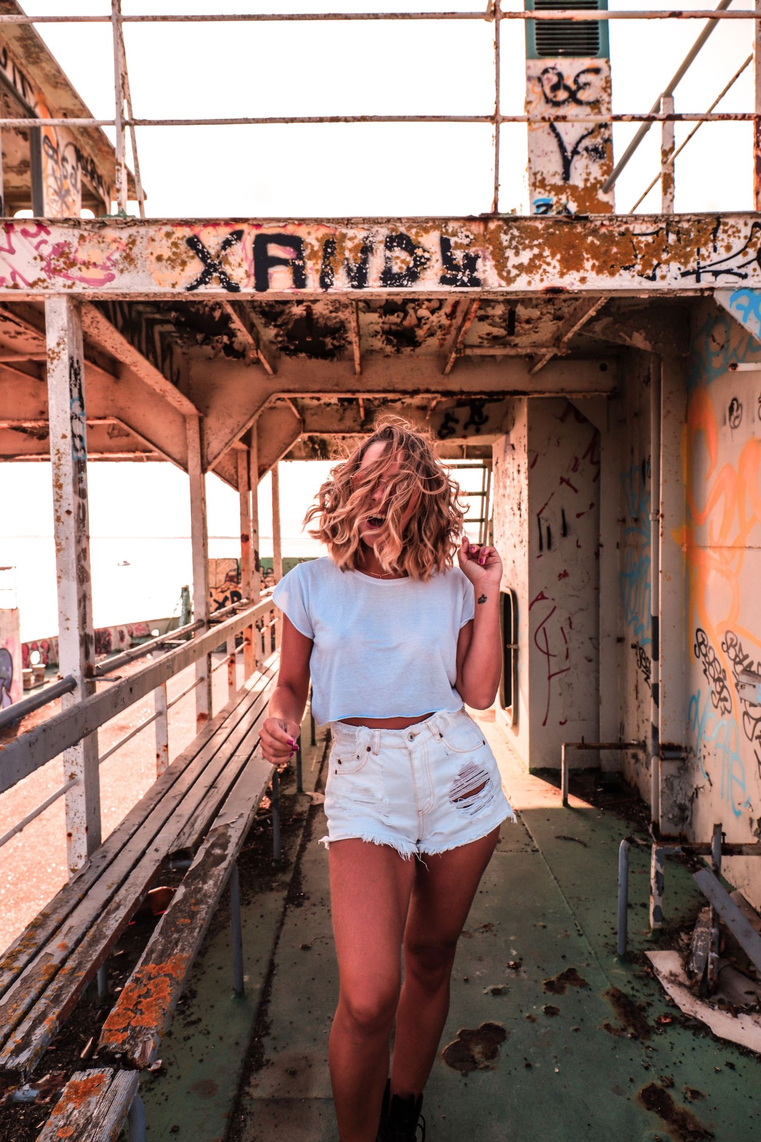 Someone in a white t-shirt and denim shorts explores a rusty abandoned industrial walkway with graffiti covered walls.