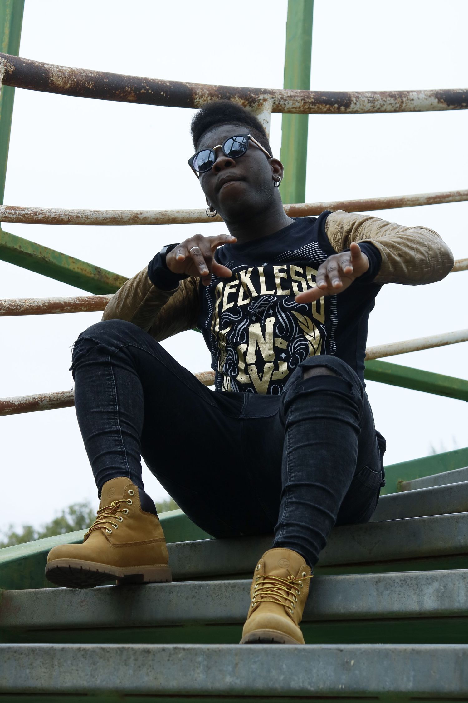 A person in a rock band t-shirt and tan boots sits casually on metal bleachers wearing sunglasses.