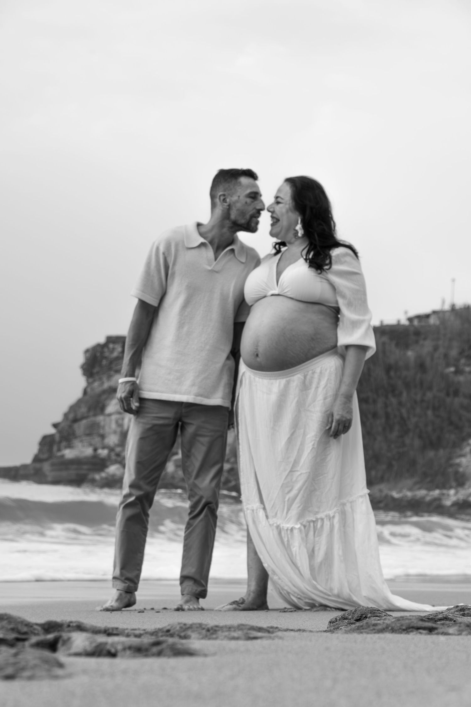 Black and white photo of a pregnant couple sharing a tender moment on a beach with rocky cliffs in the background.