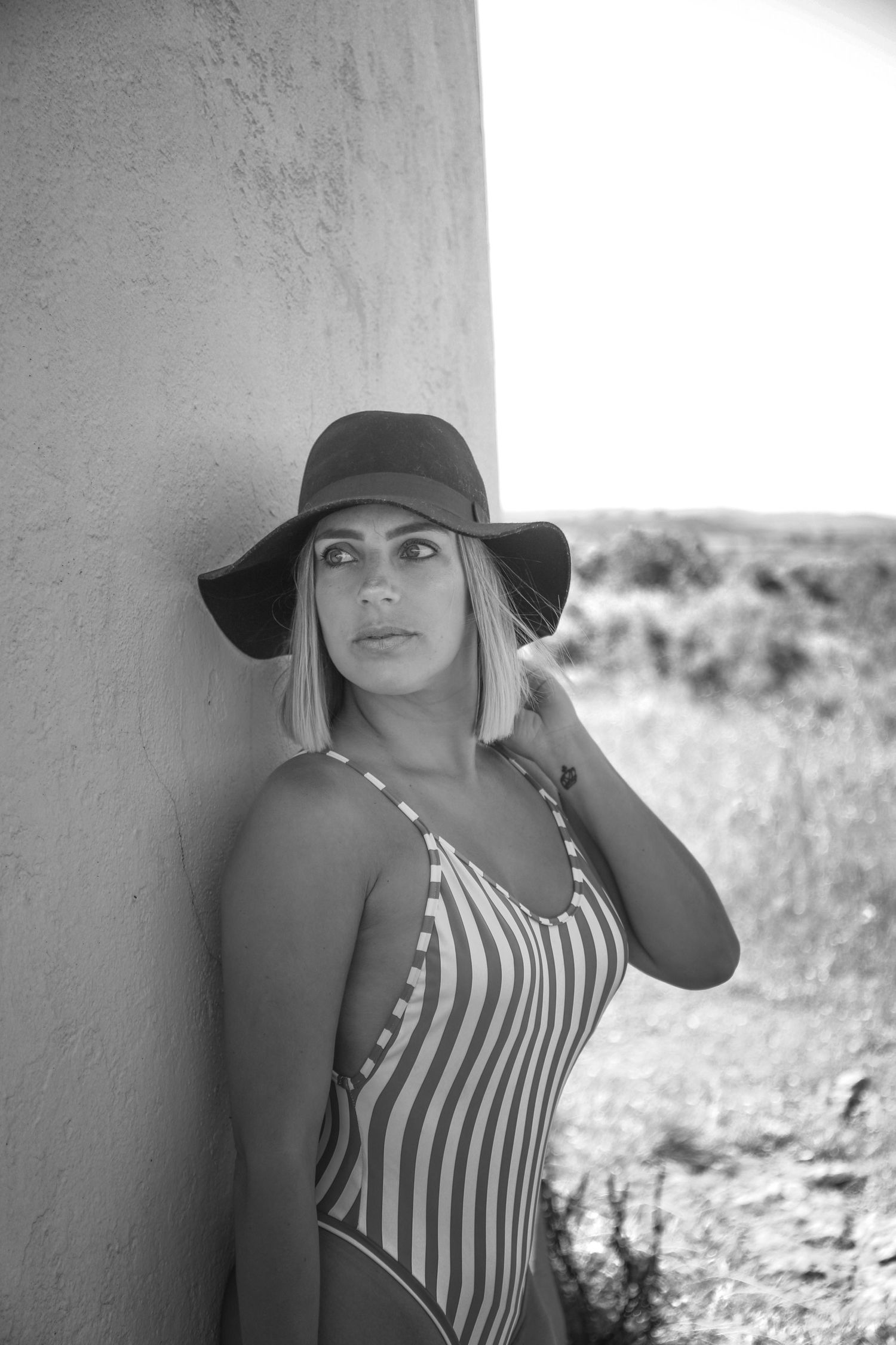 Black and white summer portrait featuring a striped swimsuit and floppy hat against a light wall outdoors.