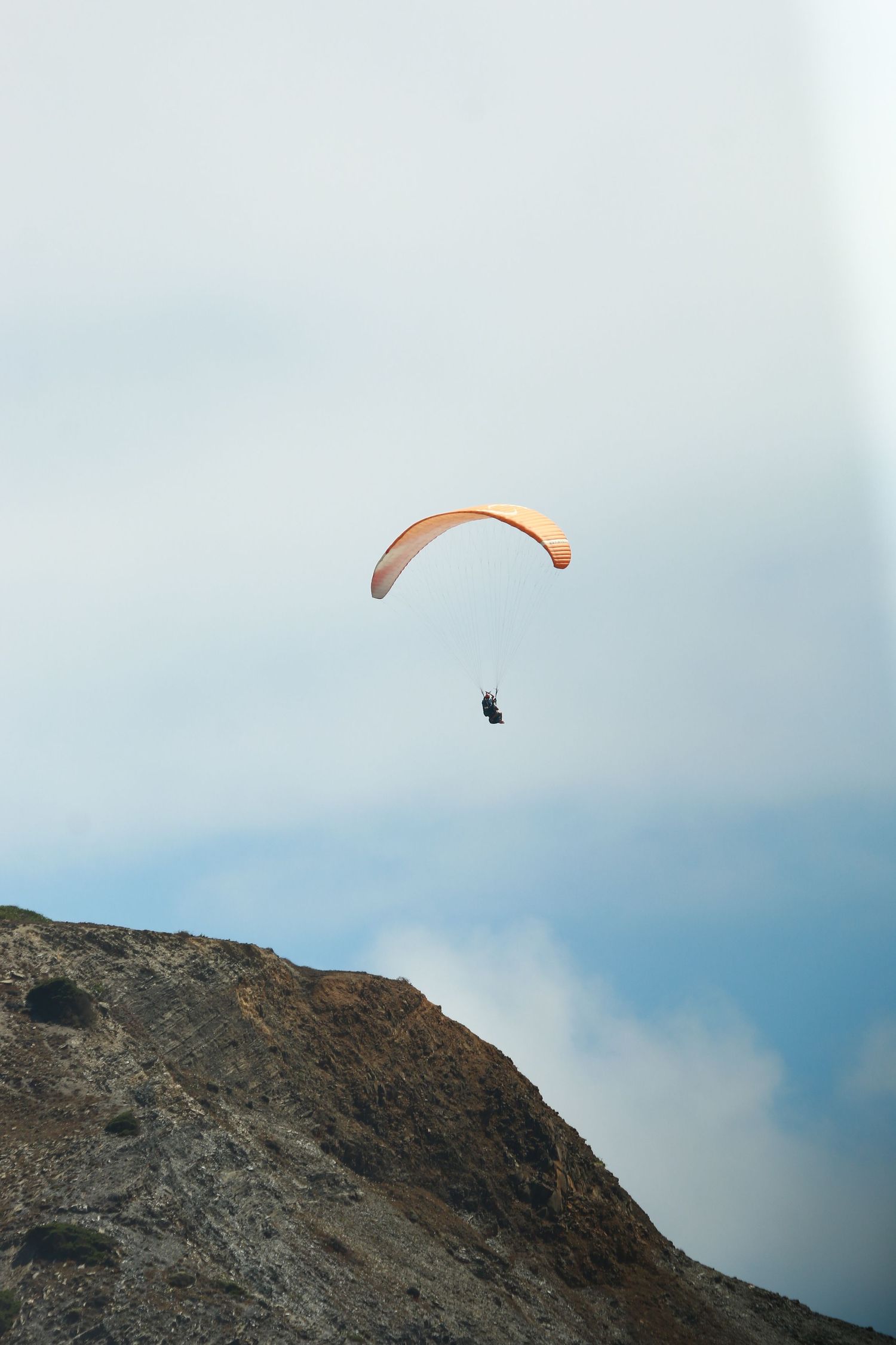 Paraglider with orange wing floating above a rugged mountainside cliff.