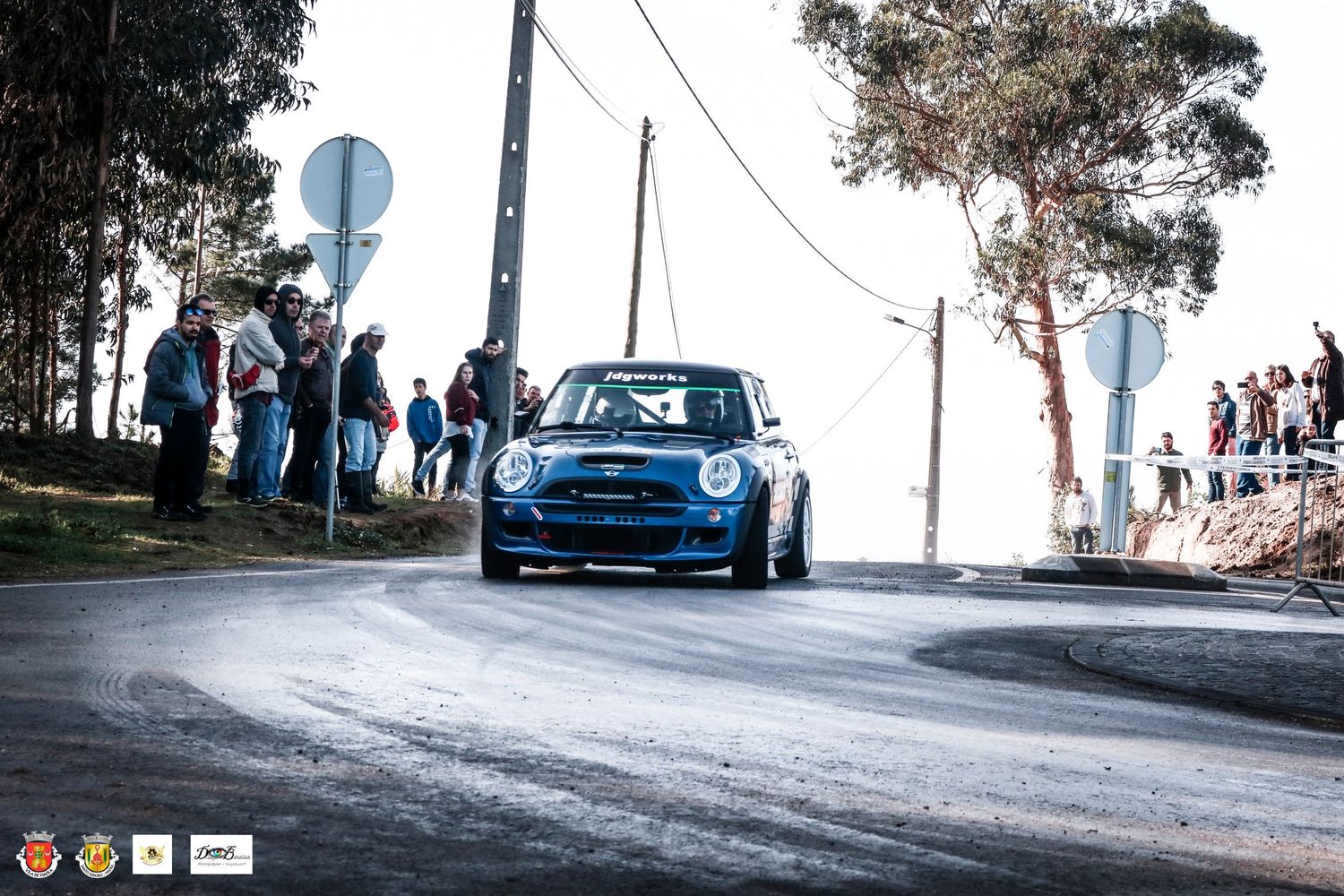 Blue classic rally car racing on wet asphalt road with spectators watching from roadside.