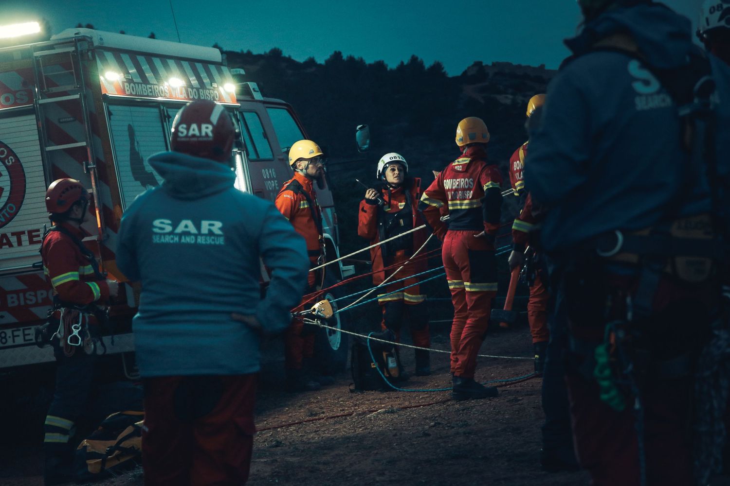 Search and rescue team in orange protective gear working at night with emergency vehicles and floodlights illuminating the scene.