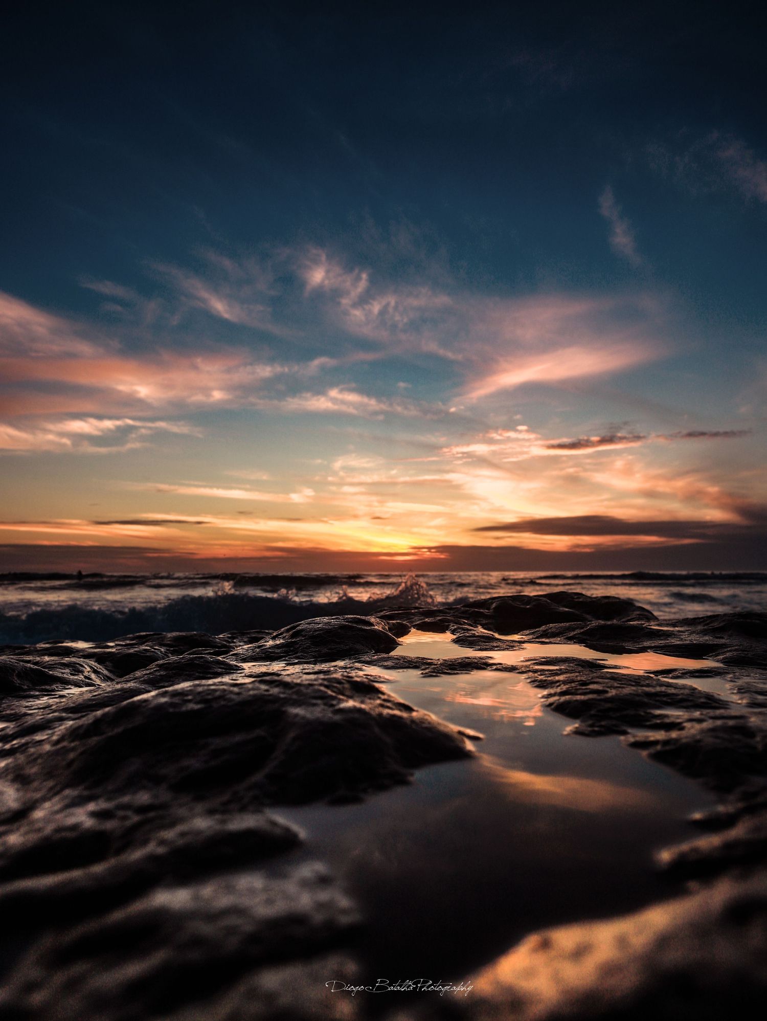 Dramatic sunset over rocky coastal tide pools with golden clouds reflecting in dark water during low tide.