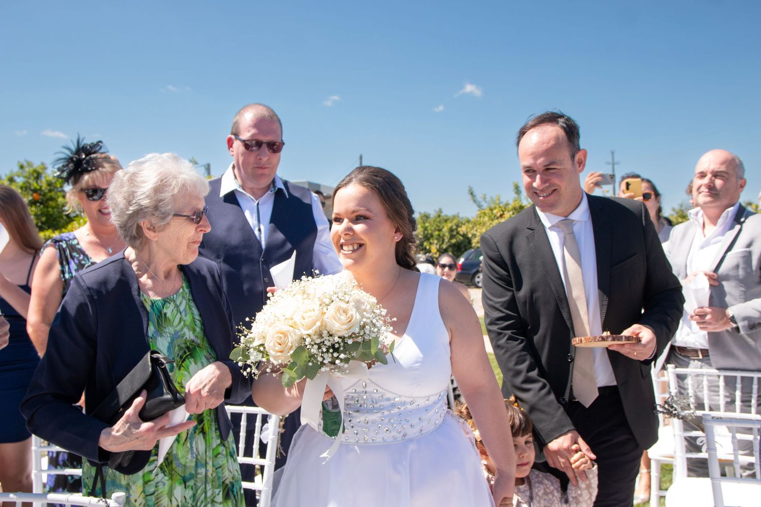 Wedding party celebrates outdoors with white chairs and bright sunshine.