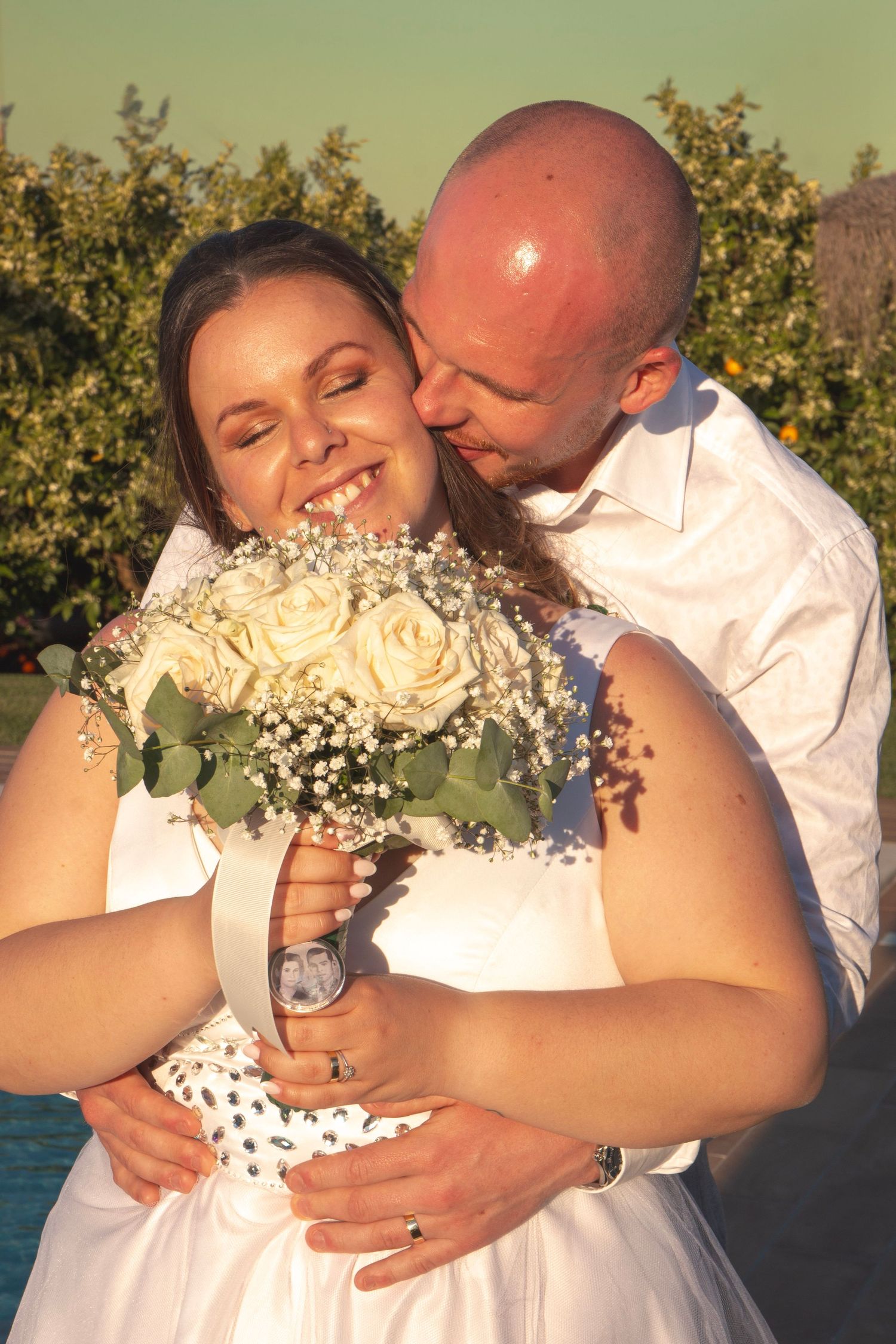 A romantic wedding moment with the couple embracing while holding a bouquet of white roses and baby's breath.