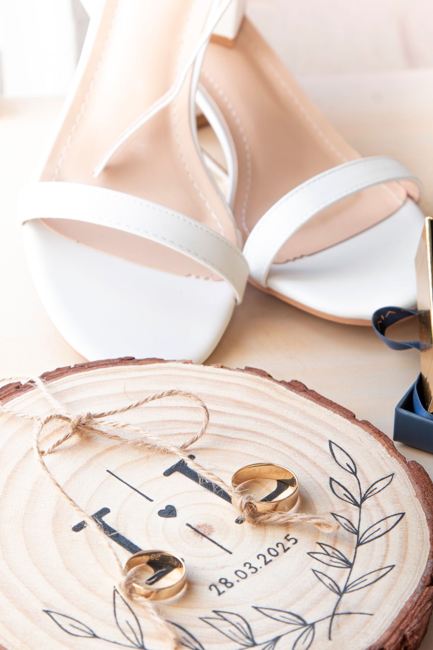 White bridal sandals and wedding rings displayed on wooden slice with date engraving.